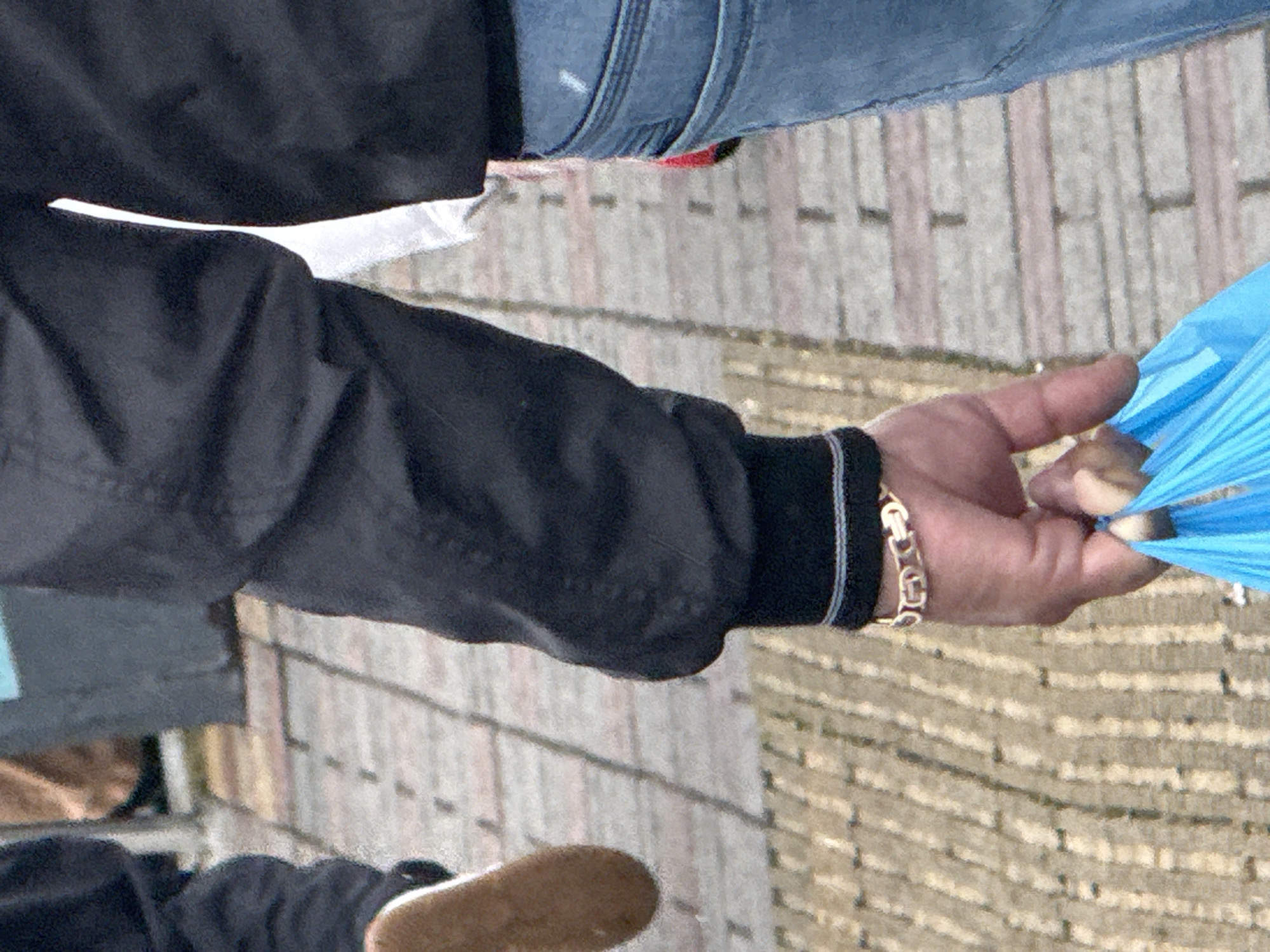A person wearing a black jacket and blue jeans stands on a brick-paved surface, possibly a walkway or street. The person's hand is visible, wearing a gold bracelet and holding a blue plastic bag. A brown shoe can also be seen. The image provides insufficient detail to confirm the location as Sochi, Russia.
