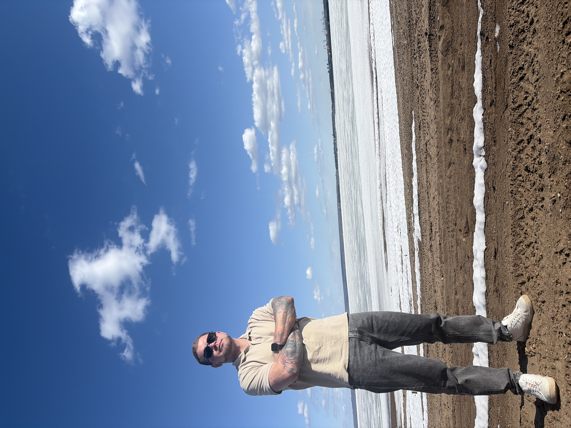 A full body shot features a man standing at the edge of a frozen lake next to the shore on a clear day with a blue sky and scattered clouds. The subject is fair-skinned, with short hair and a light beard, wearing sunglasses, a tan polo shirt, dark jeans, and white sneakers. His arms are crossed, and visible tattoos adorn both arms. The background consists of a frozen lake in the midground and a blue sky with white clouds above. The shore is on the right, marked by brown dirt and a narrow strip of snow. The location is Chaikovsky, Russia.