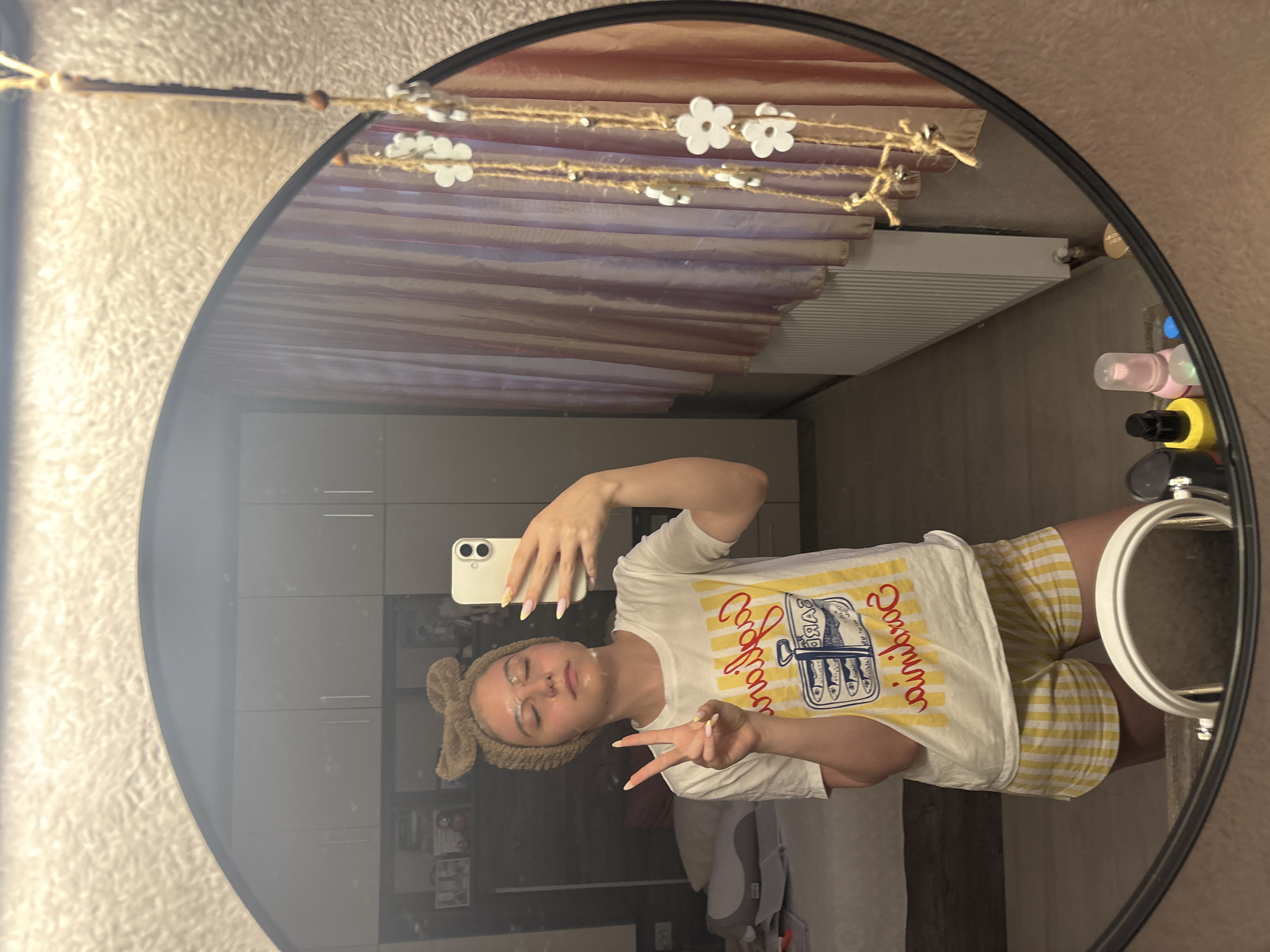 A young female is taking a selfie in a mirror inside a room. The mirror has a black frame and is decorated with lights. The female wears a white t-shirt with a graphic print, yellow and white checkered shorts, and a brown headband with a bow. Her face appears to have a skincare product on it. She is holding a white smartphone and making a peace sign with her right hand. The background includes a gray wardrobe, a radiator, and various bottles and containers on a shelf. There is no discernible information in the image to confirm it is in Bulgarovo, Bulgaria.