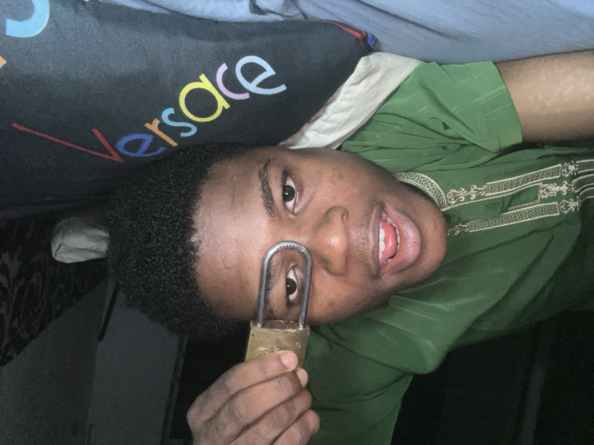 This media file shows a person lying down, with a padlock held up to their eye. The person is wearing a green garment with intricate white embroidery. Part of a black pillow with the colorful "Versace" logo is visible behind the person's head. The lighting suggests an indoor setting, possibly at night. The person's mouth is open, and their expression could be interpreted as playful or surprised. The location is identified as Ihiagwa, Nigeria.