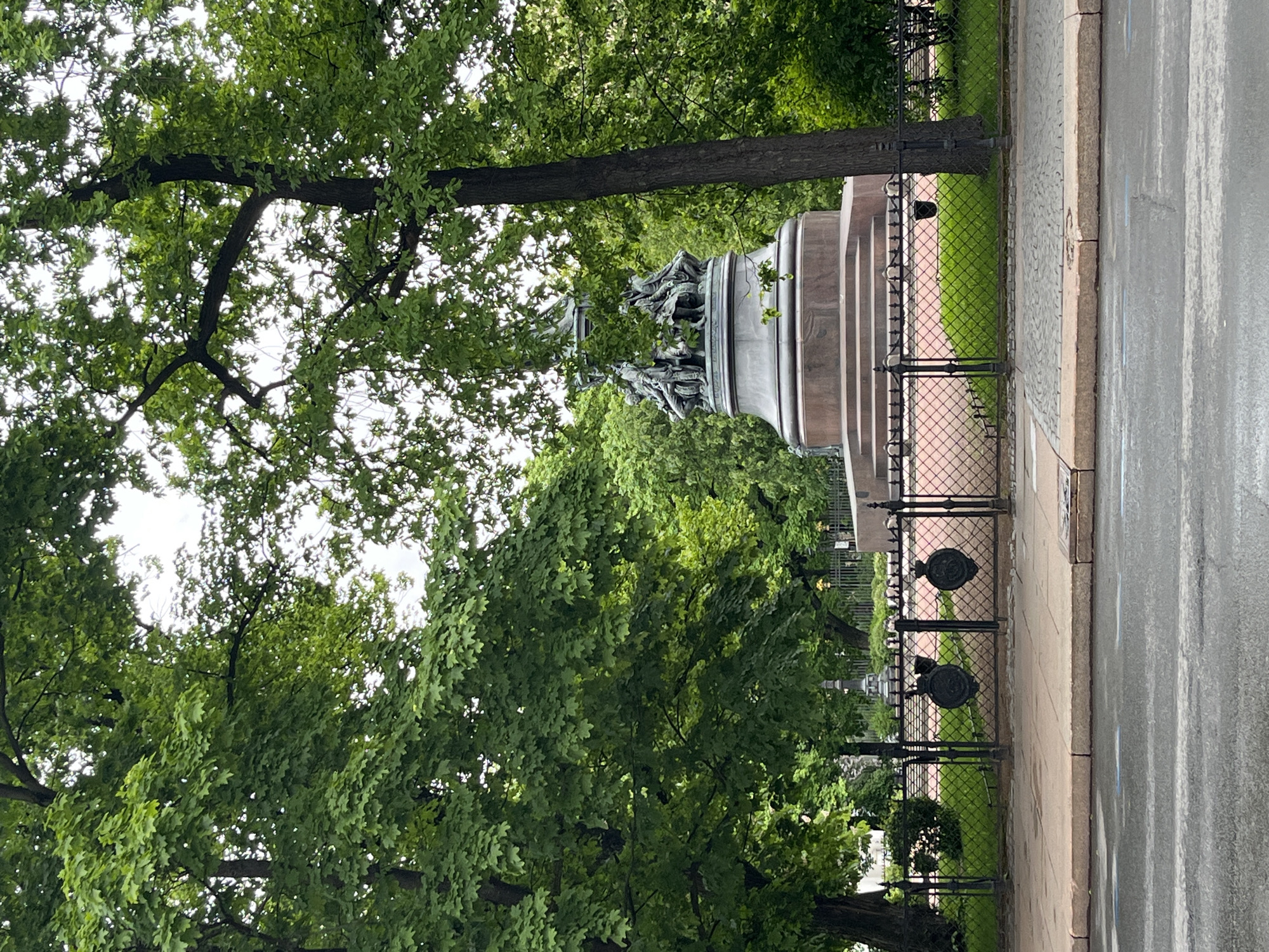 The image shows a monument in Saint Petersburg, Russia, possibly a statue or memorial, partially obscured by trees. The monument appears to be made of stone or concrete with bronze figures on top. It's surrounded by a low, ornate fence. A sidewalk runs along the edge of the park or garden where the monument is located. Beyond the sidewalk is what appears to be a street, possibly wet from recent rain. The scene is dominated by lush green trees.