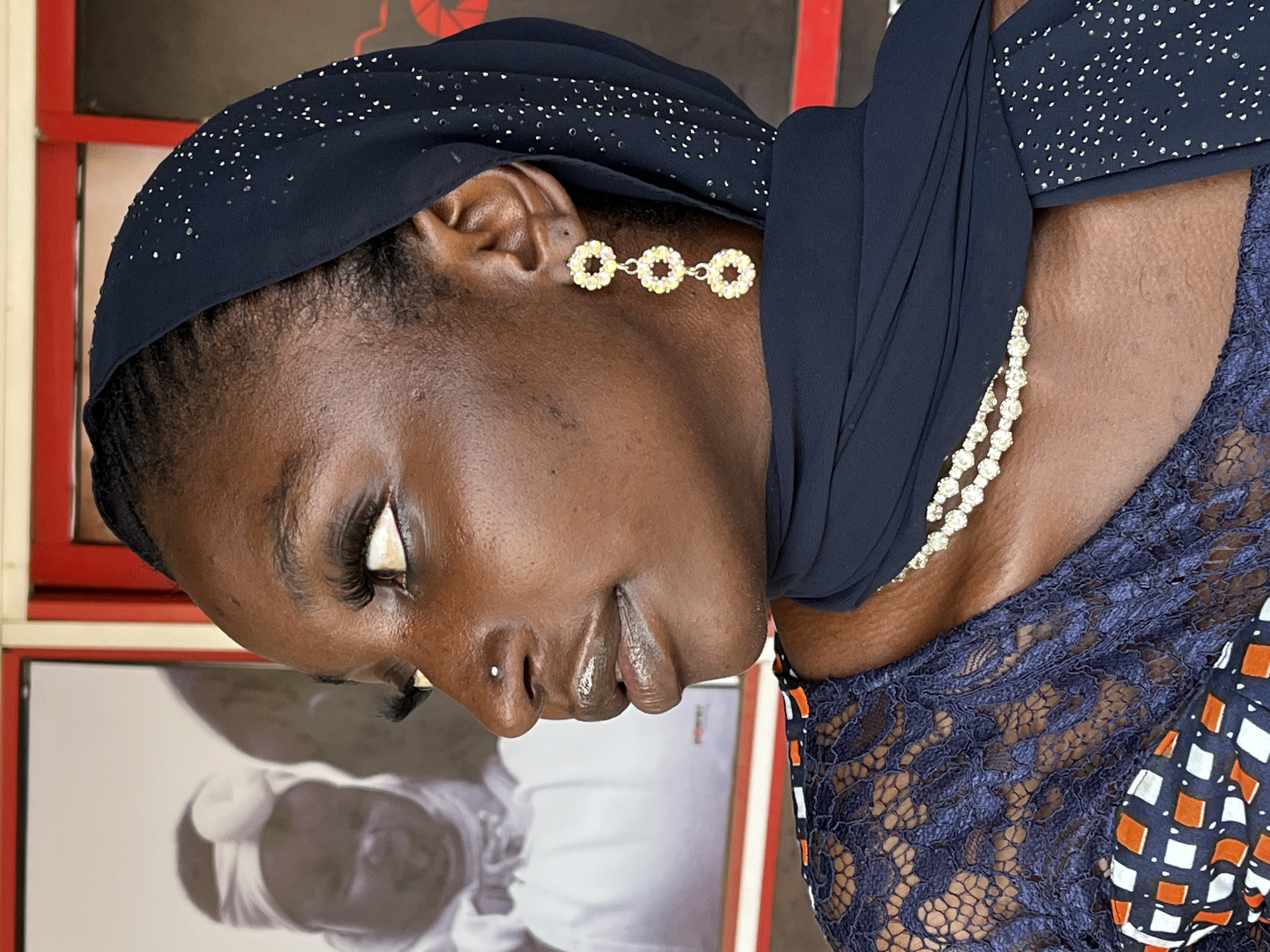A close-up, tilted portrait depicts a Black woman with brown skin wearing a dark blue head covering adorned with small, possibly reflective embellishments. A nose piercing is visible on the left nostril. She wears a short earring, visible on her right ear, made of three gold-colored circular designs. She has a two-strand necklace. The background features a vertical arrangement of framed photographs with white mattes. The background hints at a red and white framed picture, and a black and white photograph with a subject wearing a white head covering. A hint of a dark blue lace patterned garment is visible on her right shoulder. The city of Jos, Nigeria, is not visually apparent.
