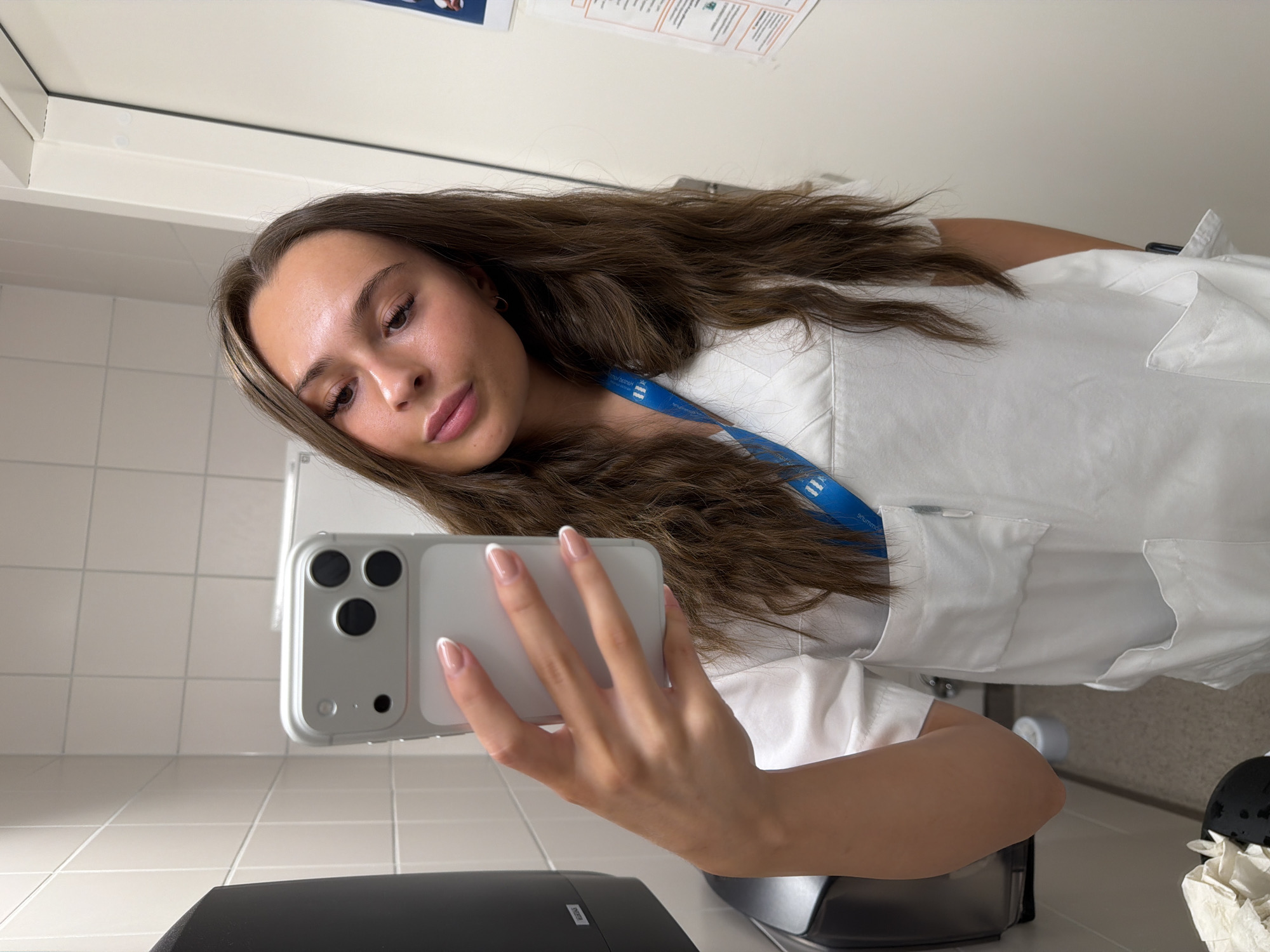 A young woman with long, wavy brown hair and fair skin is taking a mirror selfie in a brightly lit, tiled room that appears to be a restroom or changing room. She is wearing a white uniform, possibly a scrub top, with several pockets and a blue lanyard around her neck. She is holding a silver smartphone in her right hand, angled to capture her reflection. Her left arm is bent, and her hand is resting on her chest. The background shows white tiled walls, a dark dispenser on the left, and a white door or cabinet on the right. There are also some papers with text and orange borders attached to the wall on the right. The overall scene is clean and utilitarian. There is no specific visual information to indicate the location as Harstad, Norway.