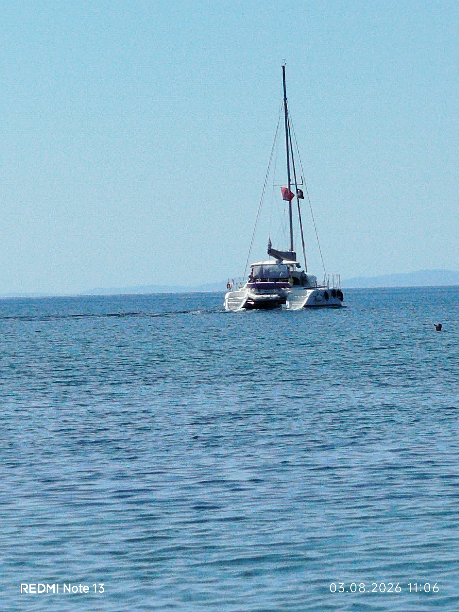 A white catamaran sailboat is navigating on a calm, blue sea under a clear, light blue sky. The catamaran has a tall mast with a red flag and a dark sail furled at the top. There are two hulls and a central cabin structure with windows and railings. Two black fenders are visible hanging from the side of the port hull. In the distance, a faint, hazy landmass is visible along the horizon. To the right of the catamaran, a person is swimming in the water, with only their head and shoulders visible. The water exhibits gentle ripples. The bottom left corner of the image displays the text "REDMI Note 13", and the bottom right corner shows the date and time "03.08.2026 11:06".