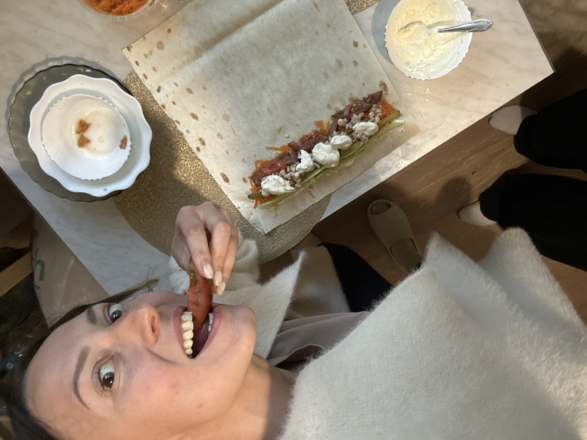 An overhead, wide-angle shot captures a woman in the process of preparing a wrap at a table. She is in the foreground, her face turned towards the camera, biting into a piece of what appears to be tomato. Her eyes are wide, and her mouth is open, revealing her teeth. She is wearing a fluffy white sweater over a pink shirt.
The table is set with a large flatbread on which ingredients for a wrap are arranged: sliced tomatoes, shredded carrots, what looks like shredded lettuce or cucumber, and dollops of white cream cheese or sour cream, topped with crumbled bits of meat or nuts. A small white bowl with a spoon contains a creamy yellow sauce, likely mayonnaise or a similar condiment. To the upper right, a clear plastic container holds more shredded carrots. A stack of two plates, one black and one white with a scalloped edge, sits to the upper right of the flatbread, with remnants of red food in the white bowl. The table itself is light-colored and appears to be made of wood or a laminate material. The floor is also visible, a dark wood grain.
The overall scene suggests a home kitchen or dining area. The presence of food preparation items and the casual attire of the woman indicate a domestic setting. The specific location of Khimki, Russia, is not visually evident from the provided image.