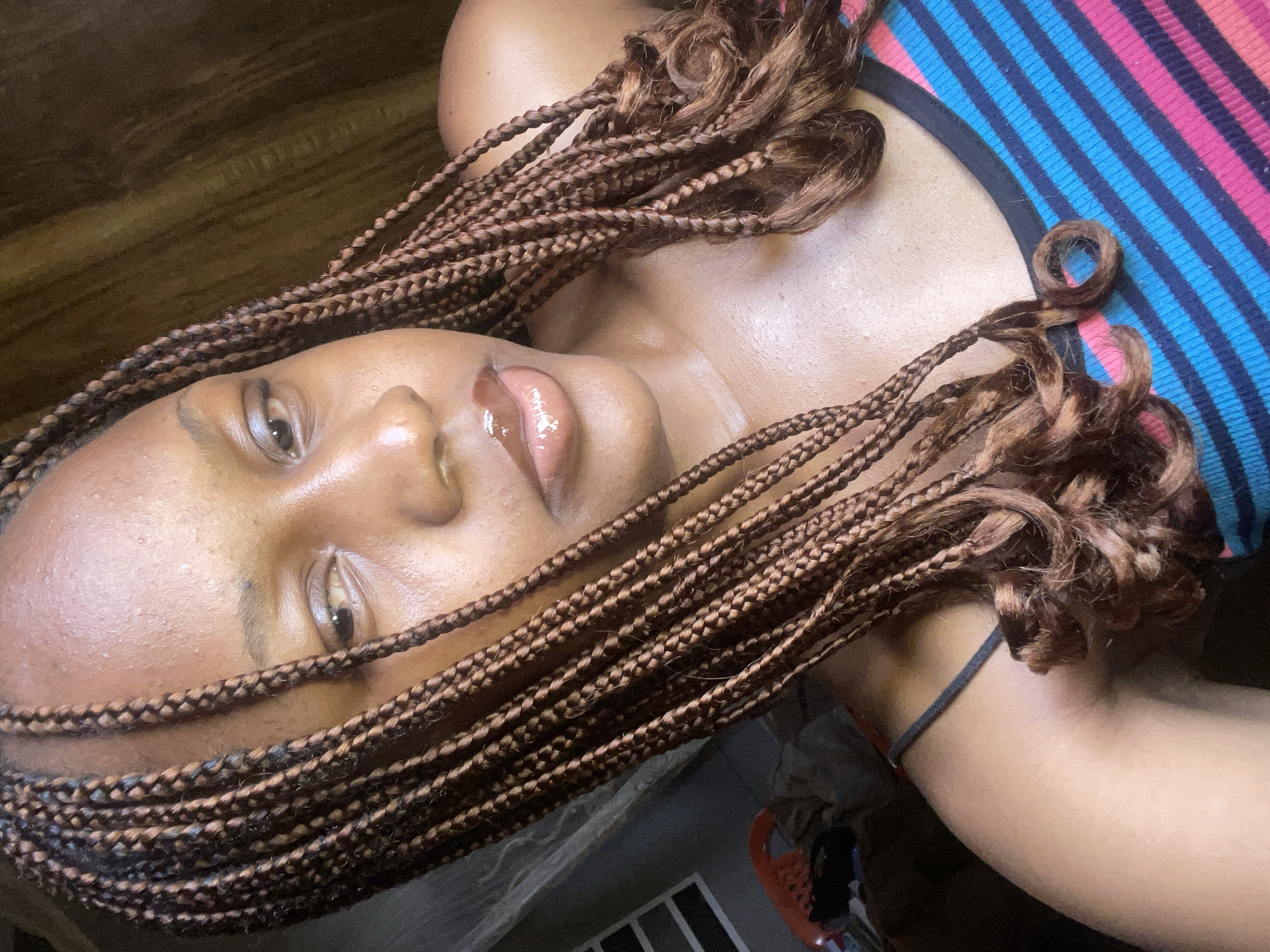 A young Black woman with braided brown hair is shown in a close-up, self-taken photograph. She is wearing a striped tank top with blue, pink, and black stripes. The background consists of a dark, textured wall and a window with blinds. There is no indication of the location being in Jos, Nigeria.