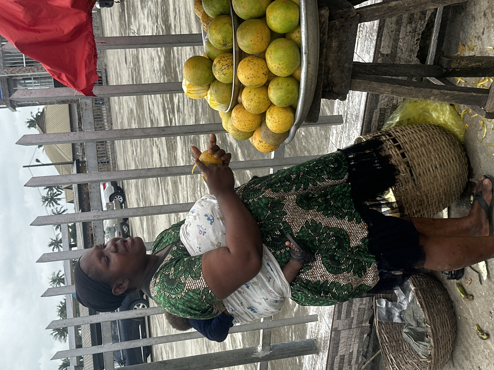 A Black woman is depicted in a medium shot, standing outside in what appears to be a market area in Badagry, Nigeria. She is wearing a green and white patterned dress and a black headscarf. A baby is strapped to her chest with a light-colored sling. The woman is holding a piece of fruit, possibly an orange, in her right hand and looking to her right. In front of her, a metal tray is piled high with oranges, with a few banana peels scattered on the ground nearby. She is standing next to a woven basket. Behind her is a metal fence and in the background, there are cars and buildings. The sky is overcast.