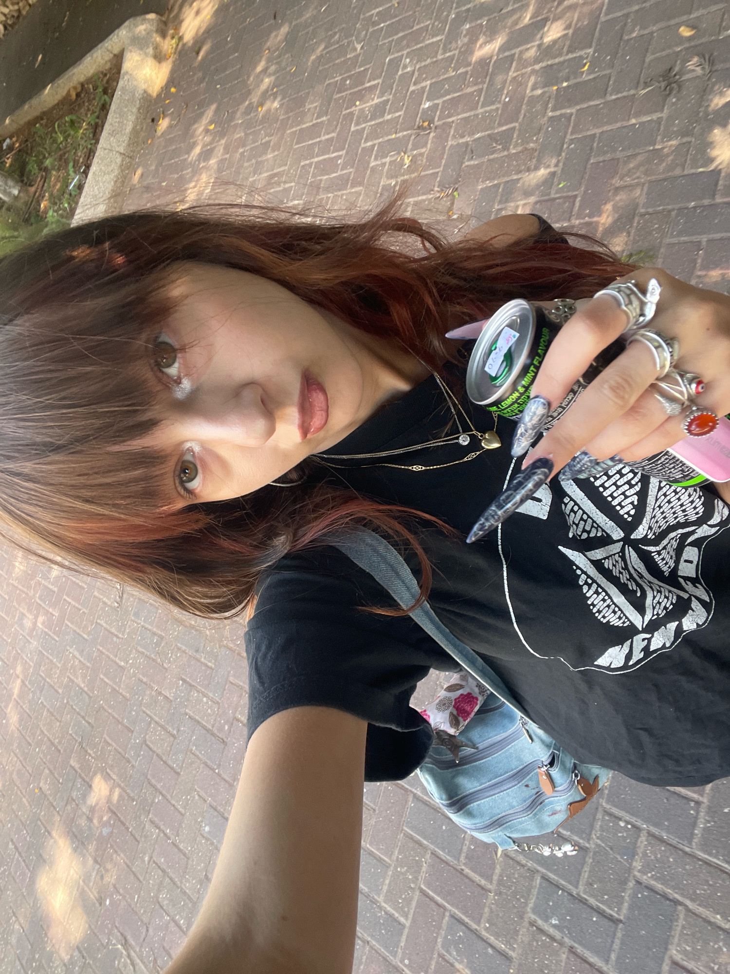 A young woman with reddish-brown hair and bangs takes a selfie in an outdoor, urban setting. She is wearing a black t-shirt with a graphic design and several rings on her fingers. She holds a can of energy drink in her right hand, which has long, pointed nails with a grey and black design. A light blue denim backpack with brown leather accents is slung over her shoulder. She is standing on a pathway paved with reddish-brown bricks, with sunlight casting shadows and highlighting parts of the ground. To the left of the pathway, there is a concrete structure and some green vegetation. The scene appears to be in Raanana, Israel, given the context of the prompt.