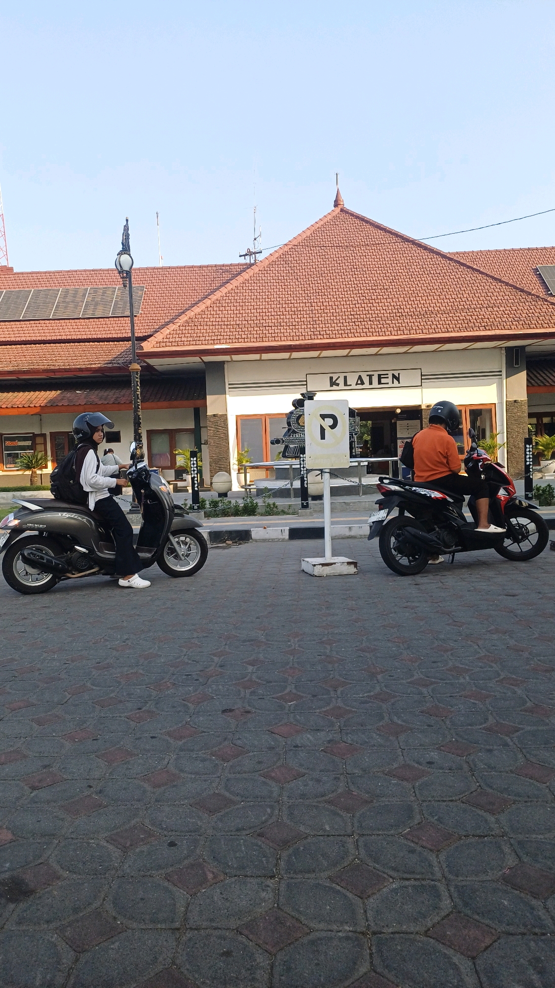 In front of a building with a sign reading "KLATEN," two individuals on scooters are captured. The building has a red tiled roof and large windows. A parking sign with a "P" symbol is visible between the two scooters. The ground is paved with dark interlocking bricks. The scooter on the left is being ridden by a person wearing a white top and black pants, with a backpack. The scooter on the right is being ridden by a person in an orange shirt and black pants. The scene appears to be an outdoor urban setting in Klaten, Indonesia, possibly near a train station given the sign. The lighting suggests it is daytime, with a clear sky.