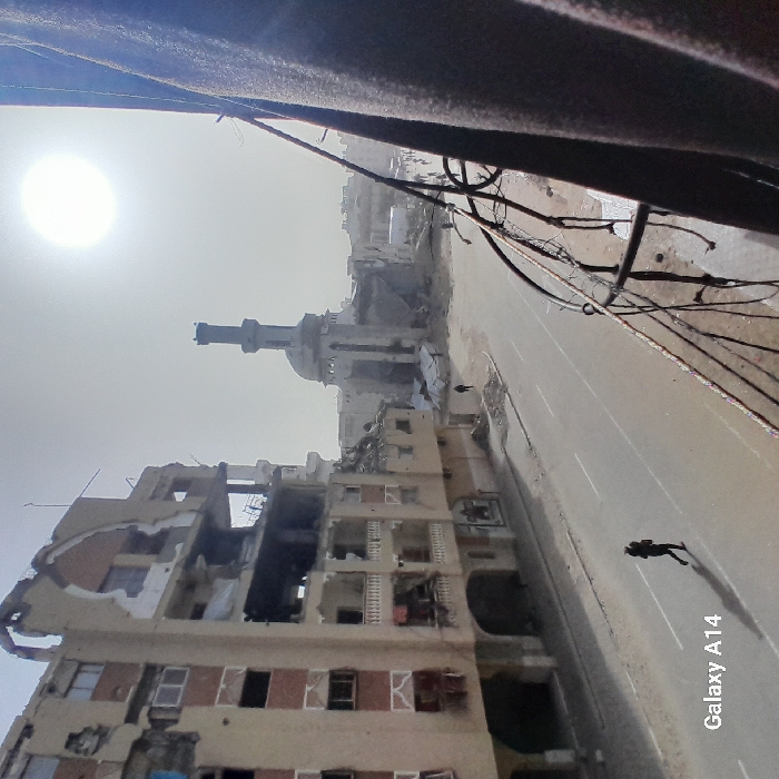 Devastation marks a street scene under a harsh sun. Buildings are severely damaged, reduced to crumbling facades and exposed interiors. A mosque's tower stands amidst the wreckage. A solitary figure walks along the empty road, contrasting sharply with the scale of destruction. Wires and debris tangle overhead, adding to the chaotic landscape. The perspective suggests the photo was taken from a high vantage point.