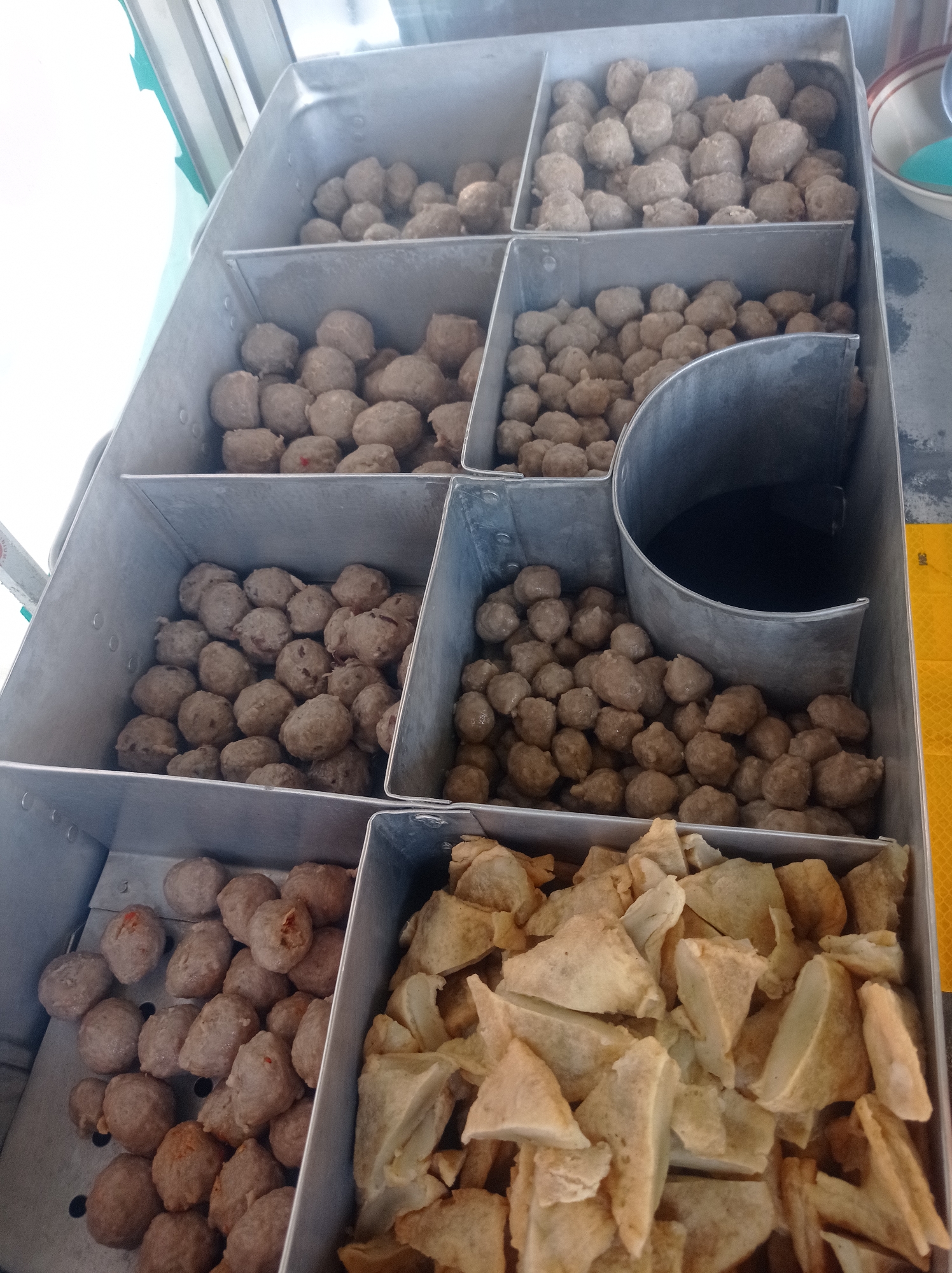 The image shows a food stall displaying several trays of food items. The stall is located in Bangunsari, Indonesia. The primary items displayed are meatballs and fried pieces of what appear to be fish cakes or tofu. The meatballs are arranged in three metal trays stacked vertically. The top two trays contain a large quantity of round, brown meatballs. The bottom left tray also contains meatballs, some of which have small red pieces visible. To the right of the meatball trays, there is a cylindrical metal container filled with a dark liquid, possibly broth or sauce. The bottom right tray contains numerous triangular, golden-brown fried pieces. The overall environment suggests a commercial food preparation or serving area, with the items likely intended for sale.