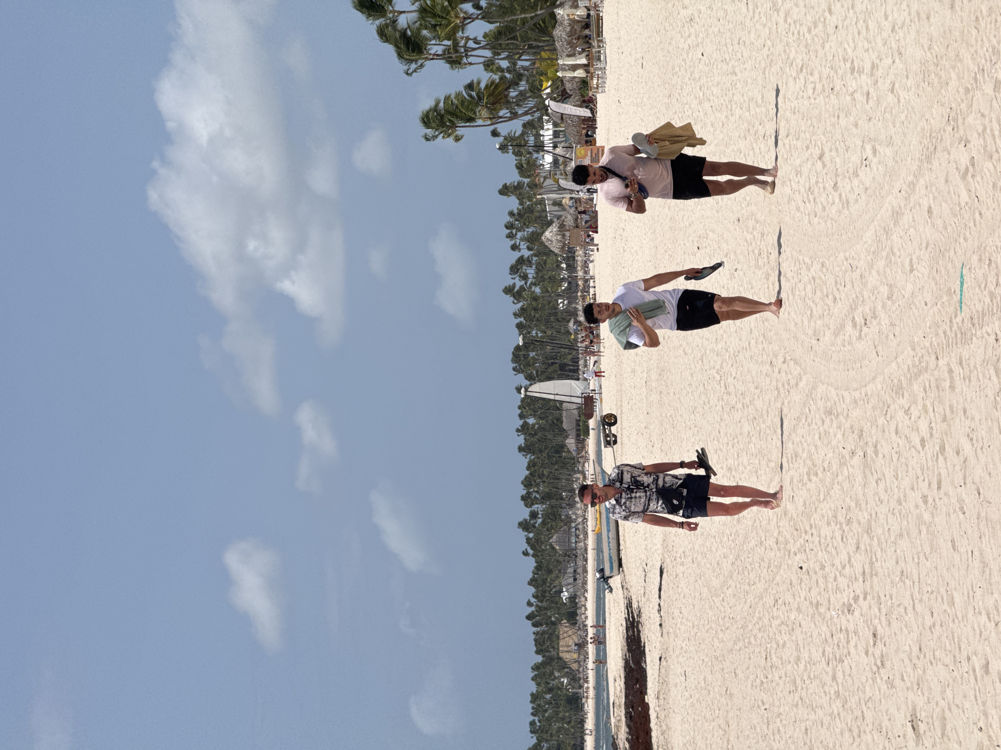 The image depicts a sunny day at a beach in Higüey, Dominican Republic. Three men are visible walking on the sandy shore, each carrying a pair of sandals. The foreground is dominated by the light-colored sand, with long shadows indicating the time of day is likely late morning or early afternoon.
In the middle ground, a line of palm trees forms a backdrop, with thatched-roof structures suggesting resort amenities. Beyond the trees, the ocean can be seen, and further in the distance, more people and beach chairs are scattered. The sky is a bright blue with scattered white clouds.
The men appear relaxed as they stroll along the beach, enjoying the tropical environment. One man in the foreground wears a patterned short-sleeved shirt and dark shorts. The man in the middle wears a white t-shirt and dark shorts. The man furthest back is wearing a pink t-shirt and dark shorts, holding a tan bag. The overall mood is one of leisure and vacation. Some visible text is present on signs in the background, though it is not legible.