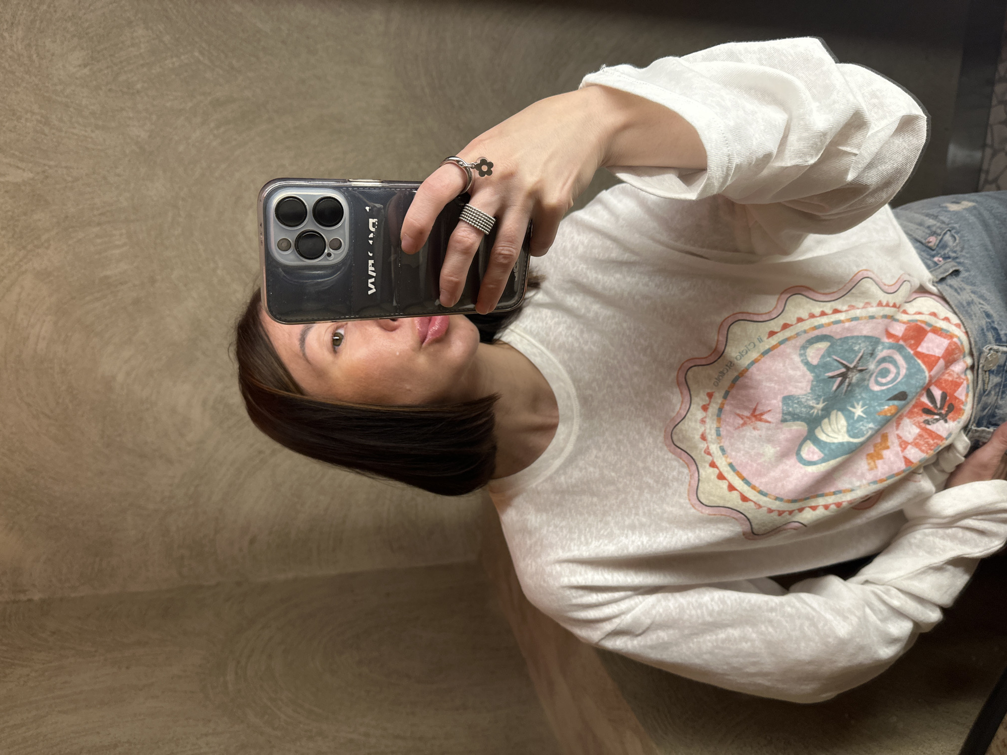 This is a mirror selfie taken indoors in the city of Усады, Russia. A woman with short, dark brown hair and lighter highlights is depicted from the chest up, holding a dark-colored smartphone with a clear case in her right hand. The phone partially obscures the right side of her face. She is looking towards the camera's reflection, with her left eye visible and her lips slightly parted, her tongue partially extended.
She wears a loose-fitting, white, long-sleeve t-shirt with a colorful, intricate graphic print centered on the chest. The graphic features an oval frame containing abstract shapes and a stylized animal figure in shades of pink, blue, orange, and red. Her lower body is partially visible, showing blue denim jeans. On her right ring finger, she wears a multi-banded silver-colored ring, and on her right pinky finger, she wears a silver-colored ring with a dangling, dark flower charm.
The background consists of textured walls in a light brownish-beige or taupe color, displaying distinct swirling patterns. A corner where two walls meet is visible on the left. A small portion of a dark, possibly tiled, floor is visible at the bottom right. The smartphone's triple camera lenses are visible on the back, and inverted text is discernible on the phone case due to the reflection.