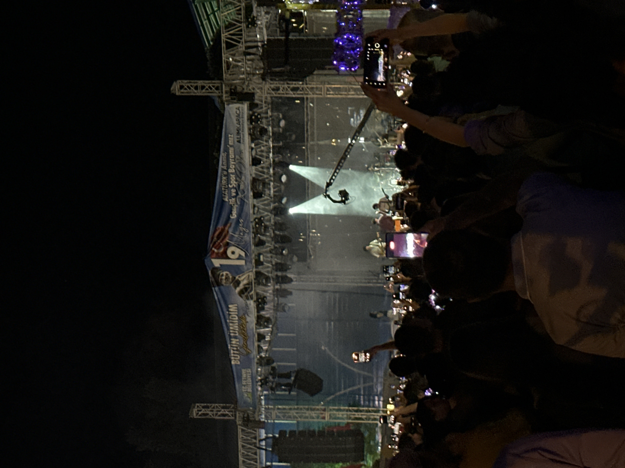 The image shows a crowd of people at an outdoor concert or event held at night in Fethiye, Turkey. The event is likely related to the commemoration of Atatürk, Youth and Sports Day. A large banner with text "BÜTÜN ÜMİDİM", the number "19", and the text "TC. FETHIYE BELEDIYESI ATATÜRK'ü Anma, Gençlik ve Spor Bayramı'mız ALIMKARACA" is displayed on the stage. People in the audience are holding up their smartphones, presumably recording or photographing the event. A stage with lighting equipment and speakers is visible.