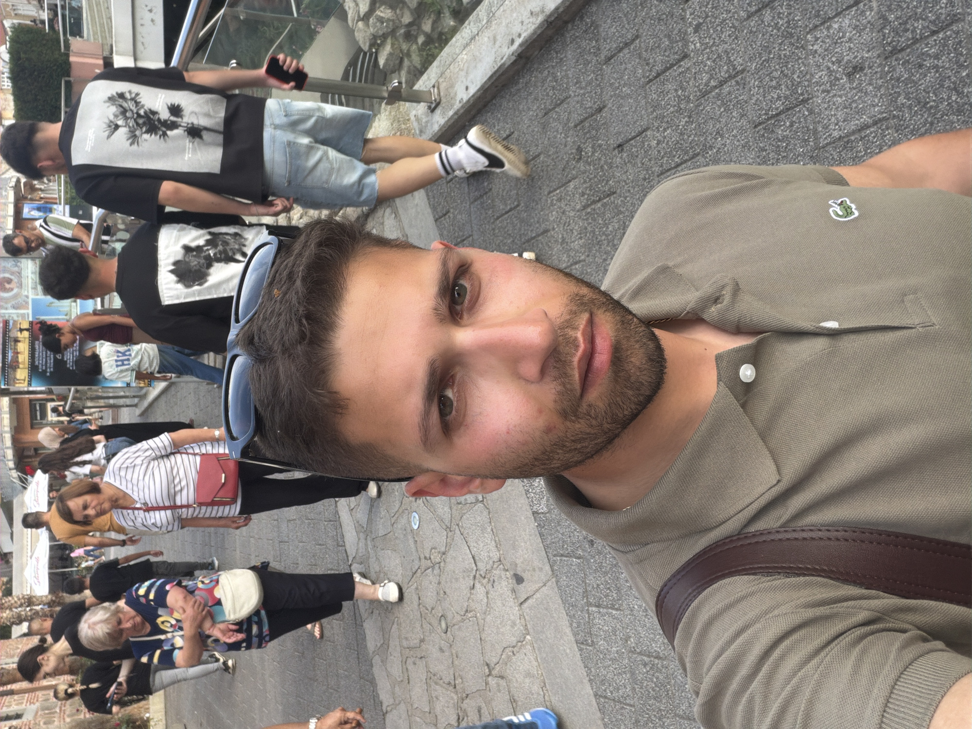 The image depicts an outdoor urban scene in Plovdiv, Bulgaria, likely a pedestrian street or promenade. A young adult male is positioned in the foreground, taking a selfie, looking directly at the camera. He has short dark hair, a trimmed beard, and is wearing an olive-green polo shirt with a white Lacoste crocodile emblem on the left chest and a brown leather strap from a bag over his right shoulder.
In the mid-ground and background, multiple individuals are walking. To the left, an older woman with short blonde hair wears a multi-colored patterned top, dark trousers, and carries a light-colored shoulder bag. Adjacent to her, another older woman with short brown hair wears a white and black striped V-neck top, dark trousers, and a red shoulder bag. Further to the right, a young adult male walks away from the camera, wearing a black T-shirt with a large white graphic of a plant/flowers on the back, light blue denim shorts, white socks, and white sneakers with black details; he holds a red smartphone in his left hand. Another male figure, similarly dressed in a black graphic T-shirt, is partially visible behind him. Other pedestrians are scattered throughout the background, including individuals near cafes with white awnings.
The ground is paved with irregular grey stone blocks, showing signs of wear. To the far right, a modern metal railing runs alongside a stone ledge, suggesting a change in elevation or an overlook. Distant buildings, characteristic of European architecture, are visible, along with some sparse vegetation including palm trees.