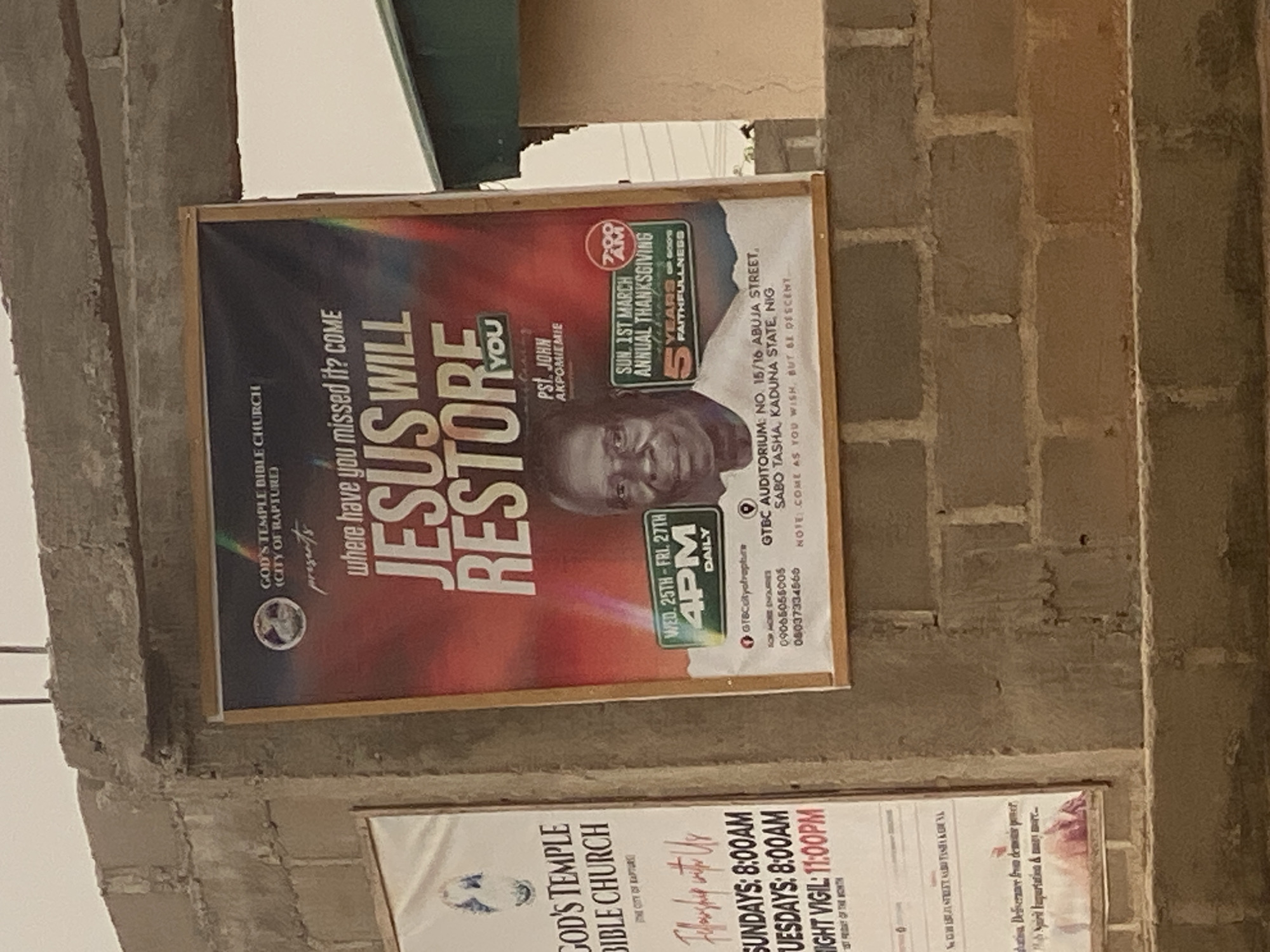 The image shows a poster for God's Temple Bible Church (City of Rapture) located at Auditorium No. 15/16 Abuja Street, Sabo Tasha, Kaduna State, Nigeria. The poster advertises a program titled "Jesus Will Restore You," presented by Pastor John Akpomiemie. It also announces the 5th annual Thanksgiving on Sunday, March 1st at 7:00 AM, with services from Wednesday the 25th to Friday the 27th at 4:00 PM daily. A photo of the pastor is featured on the poster. Another partial poster beneath it is seen, also for God's Temple Bible Church, displaying worship hours for Sundays and Tuesdays at 8:00 AM and a Night Vigil at 11:00 PM. Both posters are affixed to a brick or concrete structure.