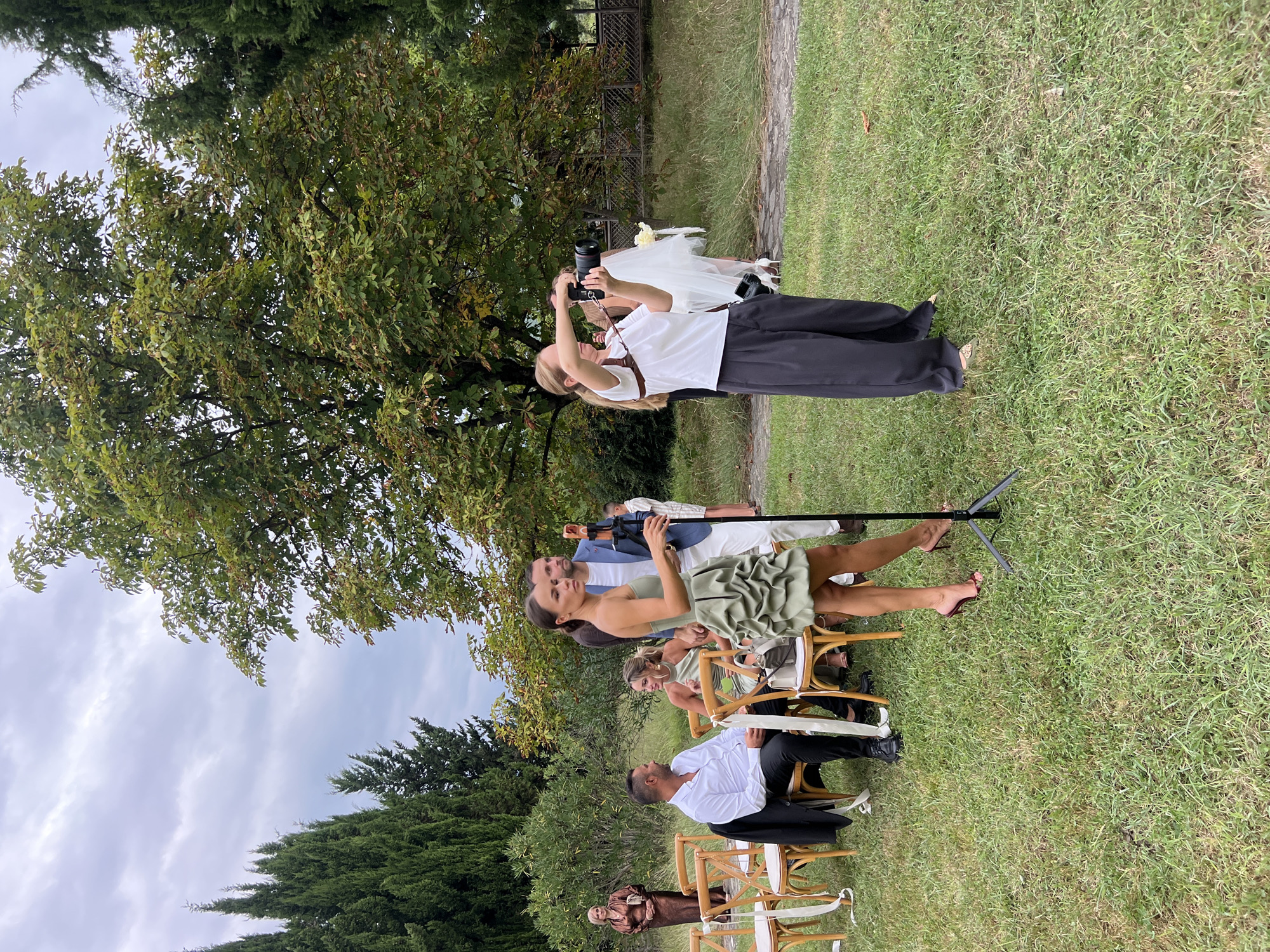 In Napareuli, Georgia, an outdoor wedding ceremony is taking place on a grassy lawn. Several guests are seated in wooden chairs arranged in rows, facing an unseen focal point. Among them is a man in a white shirt and black pants, and a woman in a green dress. To the right, a woman in a white top and dark pants is actively photographing the event with a professional camera. Another woman, dressed in a green, ruffled dress, stands near a microphone stand, appearing to be speaking or performing. Further in the background, a couple, possibly the bride and groom, are visible near a woman in a brown satin dress, with a white veil and flowers suggesting a bridal presence. The setting is surrounded by lush green trees and foliage, under a cloudy sky.