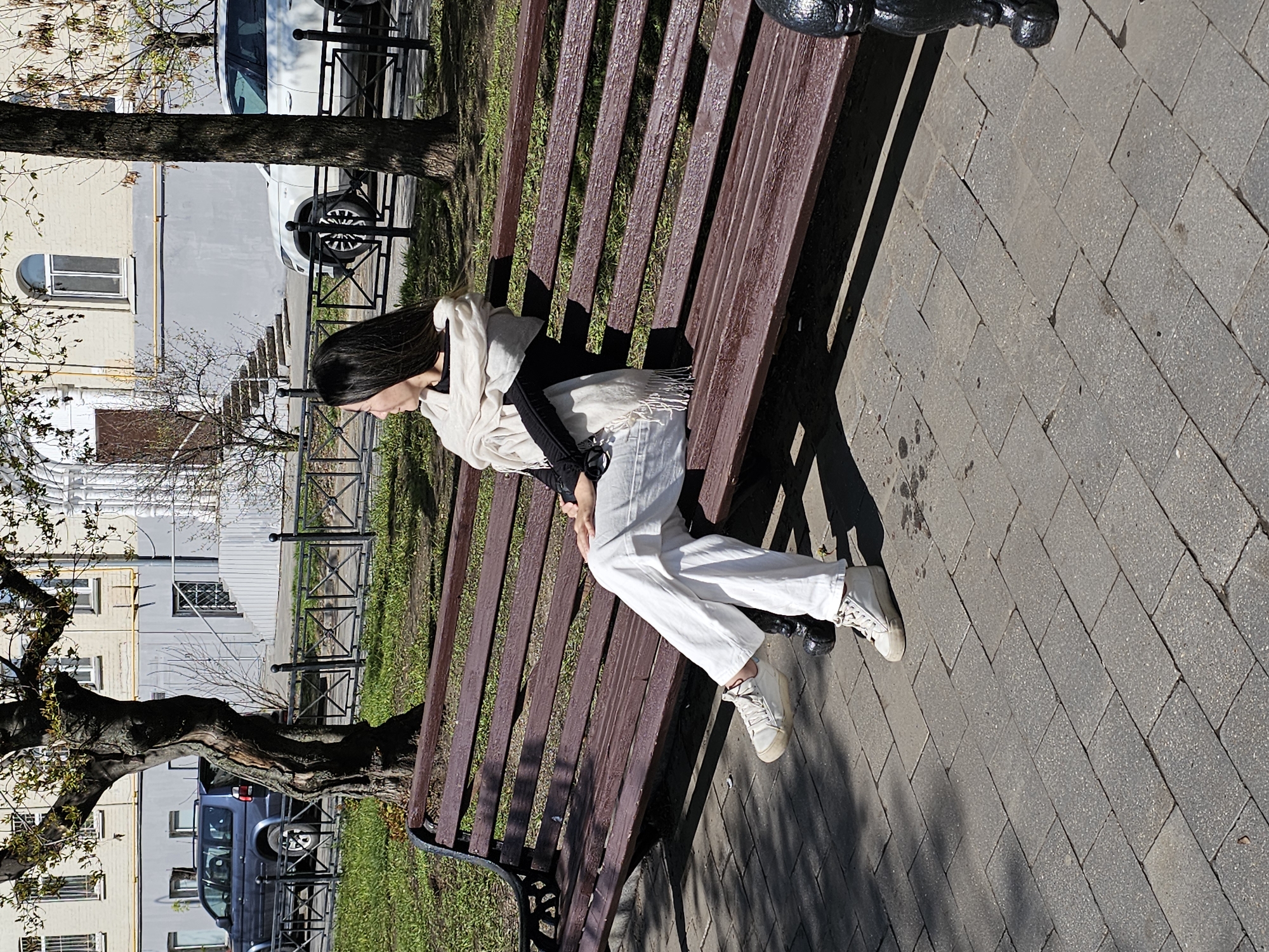 A young woman sits on a park bench in Moscow, Russia. She is fair-skinned, with long dark hair, wearing a white scarf, black top, white pants, and sneakers. The bench is made of brown slats with black metal supports, casting a shadow on the cobblestone pavement. Behind the bench is a grassy area and a fence, with buildings and trees visible in the background. Several vehicles, including a van and a car, can be seen parked near the buildings.