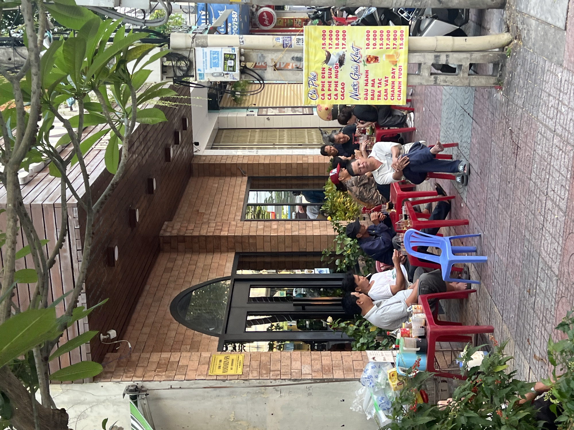 The image shows a street scene in Nha Trang, Vietnam. A group of men are seated on small plastic chairs outside a brick building. A sign advertises coffee and drinks in Vietnamese. A tree is visible on the left of the frame. Street clutter and power lines are present in the background.
