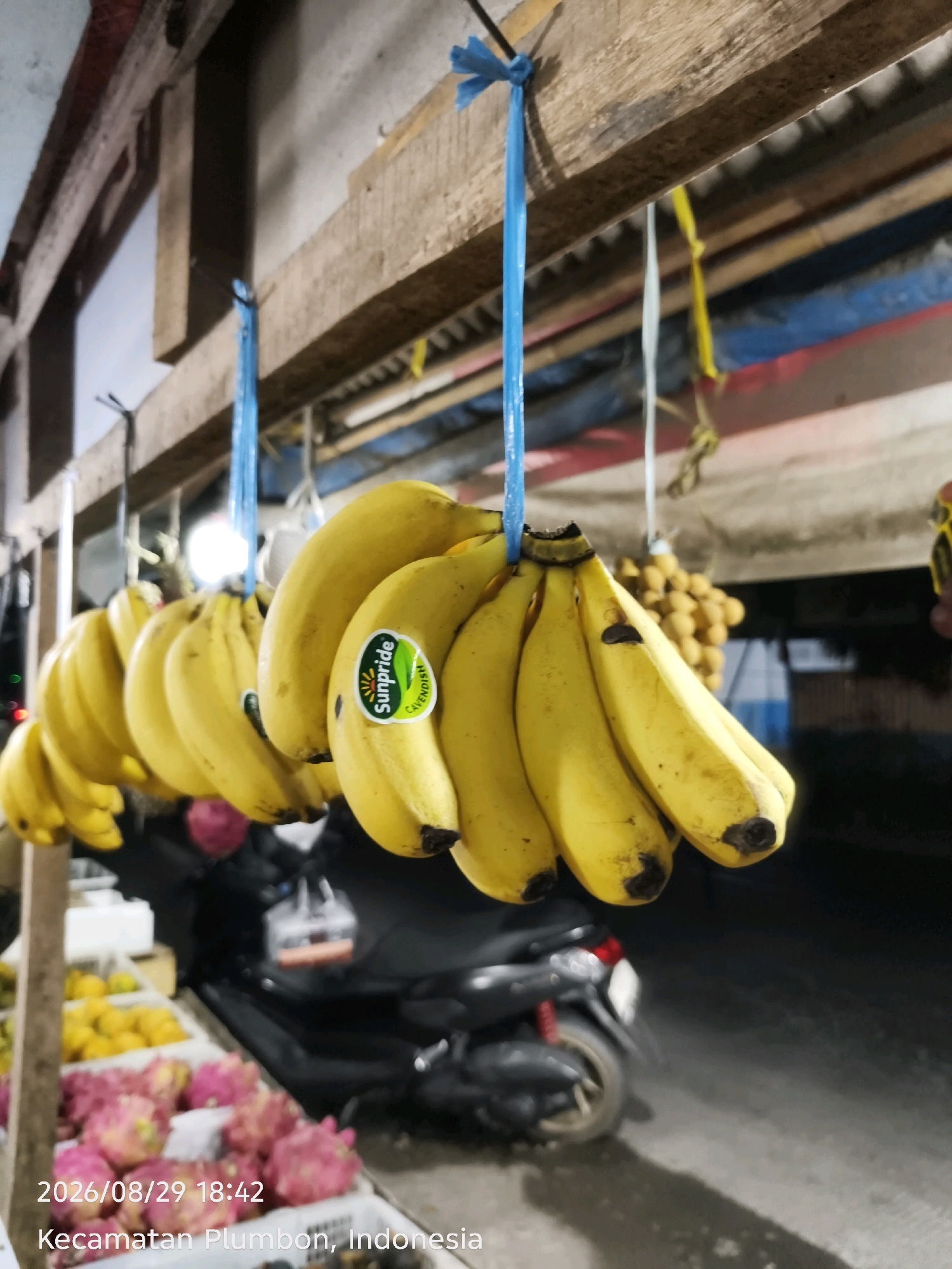 In the city of Plumbon, Indonesia, at 18:42 on August 29, 2026, a market stall displays several bunches of bright yellow bananas hanging from a wooden beam. A sticker on one bunch reads "Sunpride Cavendish." Behind the bananas, a blurry motorcycle is visible. In the foreground, trays of pink dragon fruit and yellow fruit are partially visible, along with a timestamp and location marker on the bottom left of the image. The lighting suggests it is evening.