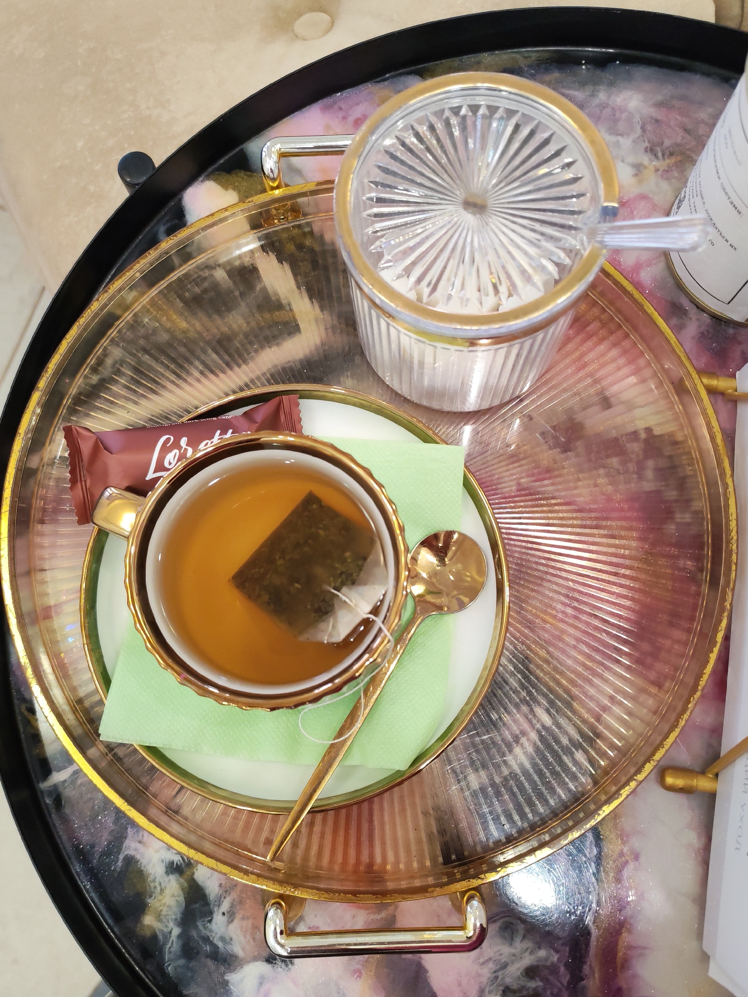 The image presents an overhead view of a single serving of tea arranged on a decorative tray. No people are visible in the scene. The setting appears to be indoors, potentially a cafe, hotel lounge, or a home in Izhevsk, Russia, given the elegant presentation and user-provided context. The lighting suggests it is daytime.
At the center of the image, a gold-rimmed white teacup holds a clear, amber-colored tea, with a teabag visibly steeping within. A small, ornate golden spoon rests beside the cup on a folded light green napkin, which sits on a white saucer. Adjacent to the teacup is a brown wrapper for a chocolate bar, prominently displaying the brand name "Loretta." Smaller, less legible text beneath "Loretta" appears to indicate "natural dark chocolate."
To the upper right of the teacup, a clear, fluted glass sugar bowl with a golden rim and a matching fluted lid is positioned. A small, clear sugar spoon extends from beneath the lid. All these items are neatly arranged on a larger, round serving tray. This primary tray features a clear, ridged or fluted glass surface encircled by a glossy gold rim. Beneath the clear glass, a striking iridescent or marbled pattern in shades of purple, pink, and white creates a decorative base. This entire ensemble rests on a larger, darker, possibly black, surface or outer tray. On the far right edge of the image, a portion of a white label or bottle is visible, displaying some Cyrillic characters such as "О", "Й", and "Н". The overall arrangement conveys a sense of readiness for a relaxing tea break.