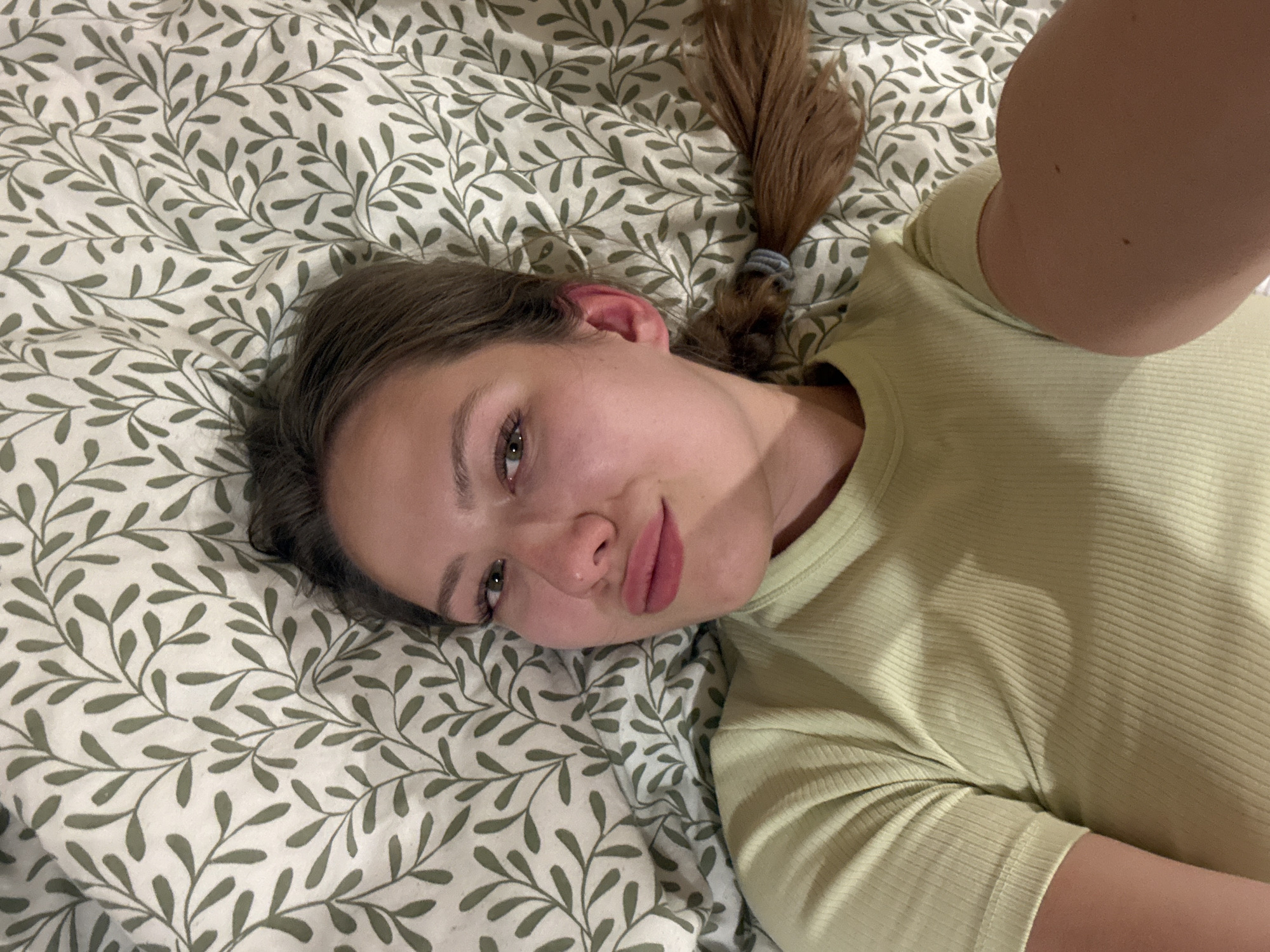 A young woman with light brown hair, styled in a braid, is lying on a bed with a white and green leaf-patterned duvet cover. She's wearing a light green ribbed t-shirt and looking at the camera. The image appears to be a selfie taken indoors, presumably in a bedroom. The location is not provided in the image.