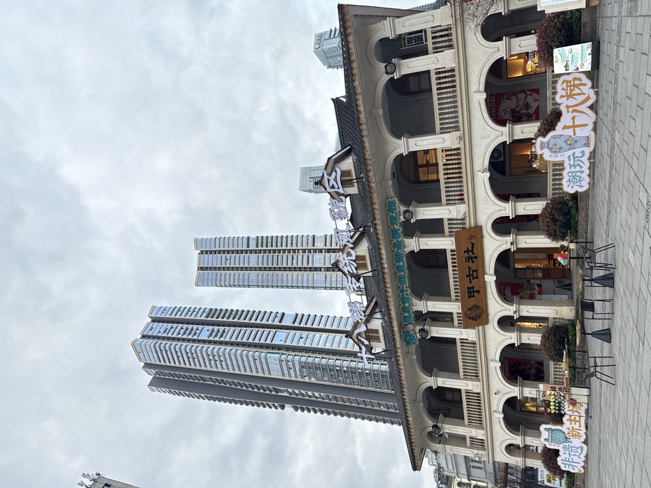 The image presents a street-level view of the Shibatizi Traditional Style Area in Yuzhong District, Chongqing, China, on an overcast day. In the foreground stands a two-story building of traditional Chinese design, featuring light-colored walls, a dark tiled roof with three dormer windows, and a continuous balcony on the second floor supported by a series of arches and a balustrade. The ground floor mirrors this with a colonnade of arches, revealing illuminated shop interiors. A wooden sign displaying "甲古社" (Jiagushi) is centrally mounted above the ground floor arches, while larger characters spelling "十八梯传统风貌区" (Shibatizi Traditional Style Area) are positioned on the roofline.
Behind and towering over this traditional structure are two prominent, modern skyscrapers with extensive glass facades, along with other high-rise buildings partially visible in the distant background.
The paved plaza in front of the traditional building is furnished with four dark-colored round tables on conical bases and several matching chairs, arranged for outdoor seating. Two large, white promotional cutouts flank the seating area: one on the left featuring a light blue, pot-shaped cartoon character with text "非遗 新主场" (Intangible Cultural Heritage New Main Venue) and a display of colorful items; the other on the right depicts a white vase with blue patterns and text "潮玩 十八梯" (Trendy Play Shibatizi). Small branches with red blossoms are visible on the far right. No prominent individuals or discernible actions are present in the immediate scene.