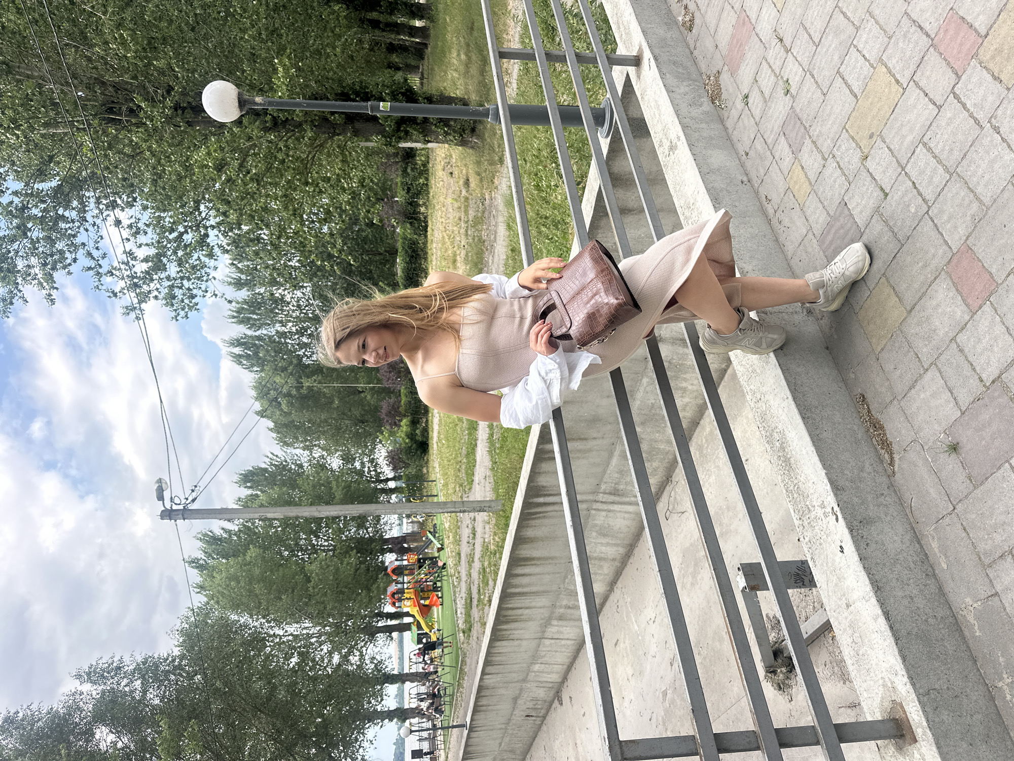 A young woman is standing on a paved walkway next to a concrete barrier and a metal railing. She is wearing a light pink slip dress with a white button-down shirt draped over her shoulders, and light gray sneakers. She is holding a brown alligator-print handbag. The background shows a playground with colorful equipment, trees, and a body of water. The sky is partly cloudy. This scene is located near the Ternopil Pond in Ternopil, Ukraine.