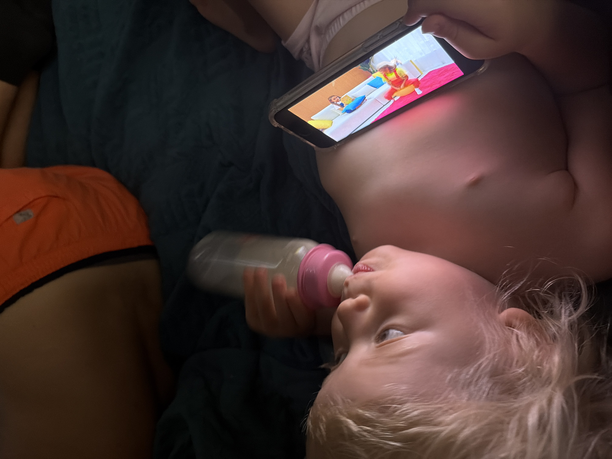 The media file shows two young children lying down. One child, in the foreground, is a blonde toddler with light blue eyes. They are holding a baby bottle with a pink nipple and drinking from it. Their attention is focused on a smartphone held by someone off-camera, which displays a bright, colorful scene featuring animated characters. The second child is partially visible on the left side of the frame, wearing orange shorts with a black waistband. They are lying on a dark blue, textured surface, possibly a blanket or bed. The scene appears to be indoors, in a dimly lit room. The overall impression is one of quiet relaxation and engagement with digital entertainment. There is no visible text in the media file.