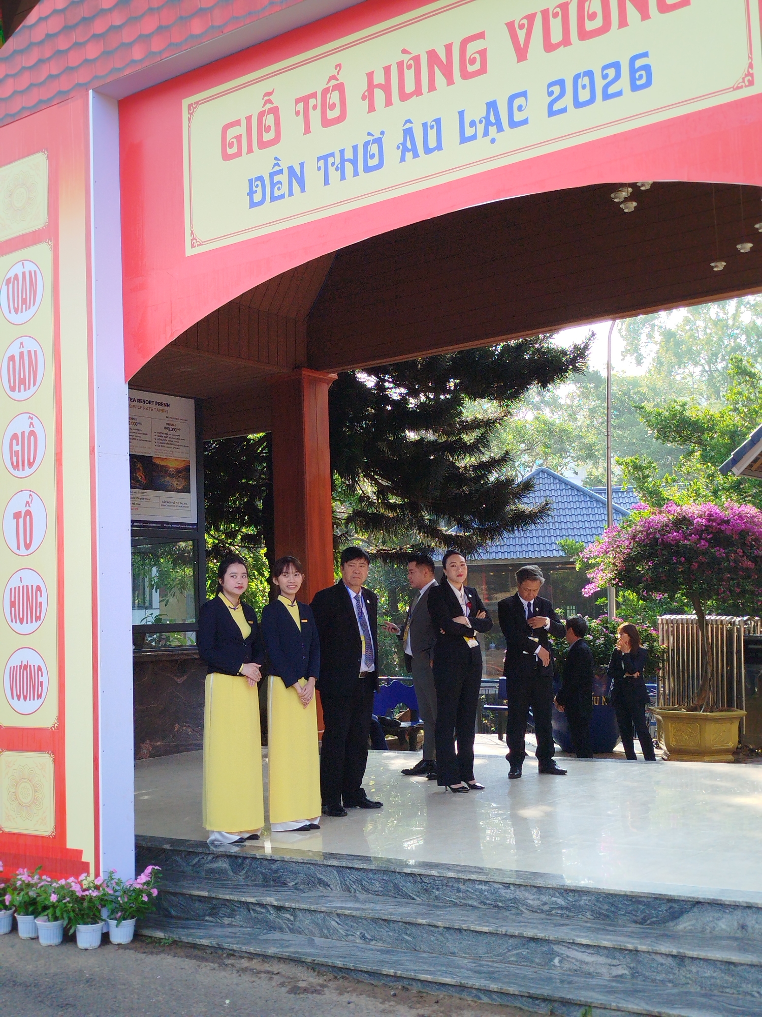 The image shows a group of people standing on a platform, likely a stage or entrance area, in what appears to be a public space. Two women stand on the left wearing traditional Vietnamese dresses (Áo dài) in yellow with navy blue jackets. A man in a suit stands next to them, also facing forward. Other individuals are seen further back, also dressed in formal attire, some engaged in conversation. A sign above the entrance reads "GIỖ TỔ HÙNG VƯƠNG ĐỀN THỜ ÂU LẠC 2026", suggesting a memorial event for the Hung Kings, taking place in 2026, at a location named Âu Lạc. A service rate tariff board is visible near the women. A structure with a red and yellow frame acts as an entrance or gateway. The scene includes plants, a building with a blue roof visible in the background, and trees, possibly in Da Lat, Vietnam.
