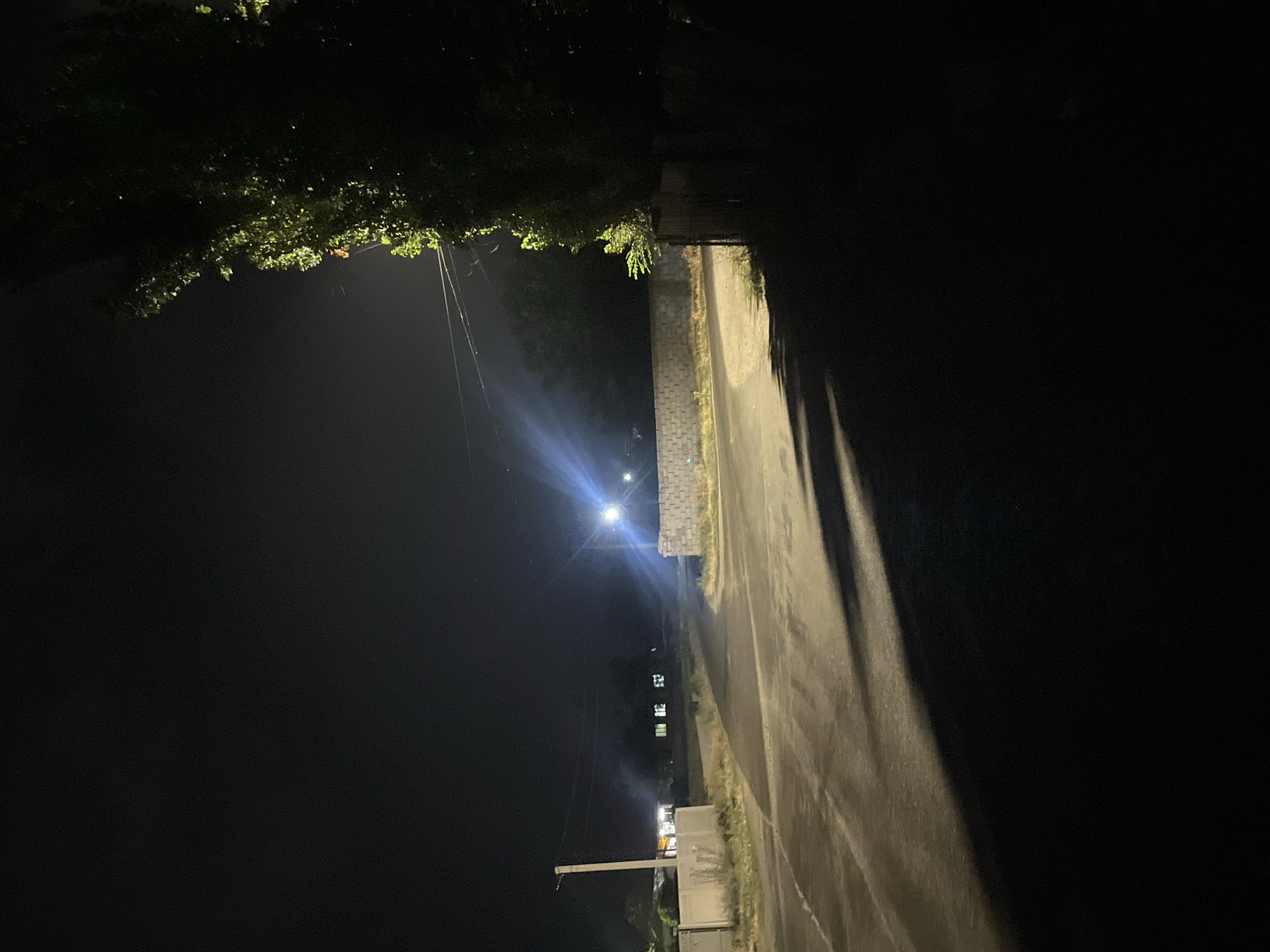 The image depicts a nighttime scene on a paved road in the city of Ak-Jol, Kyrgyzstan. A street light illuminates a portion of the road, casting long shadows. To the right, a large tree with green foliage is visible, its leaves catching the light. A low, grey concrete wall runs along the side of the road. In the background, houses with illuminated windows can be seen, suggesting a residential area. Power lines are strung across the scene, supported by a utility pole on the left. The overall atmosphere is dark and quiet, characteristic of a rural or suburban setting at night.