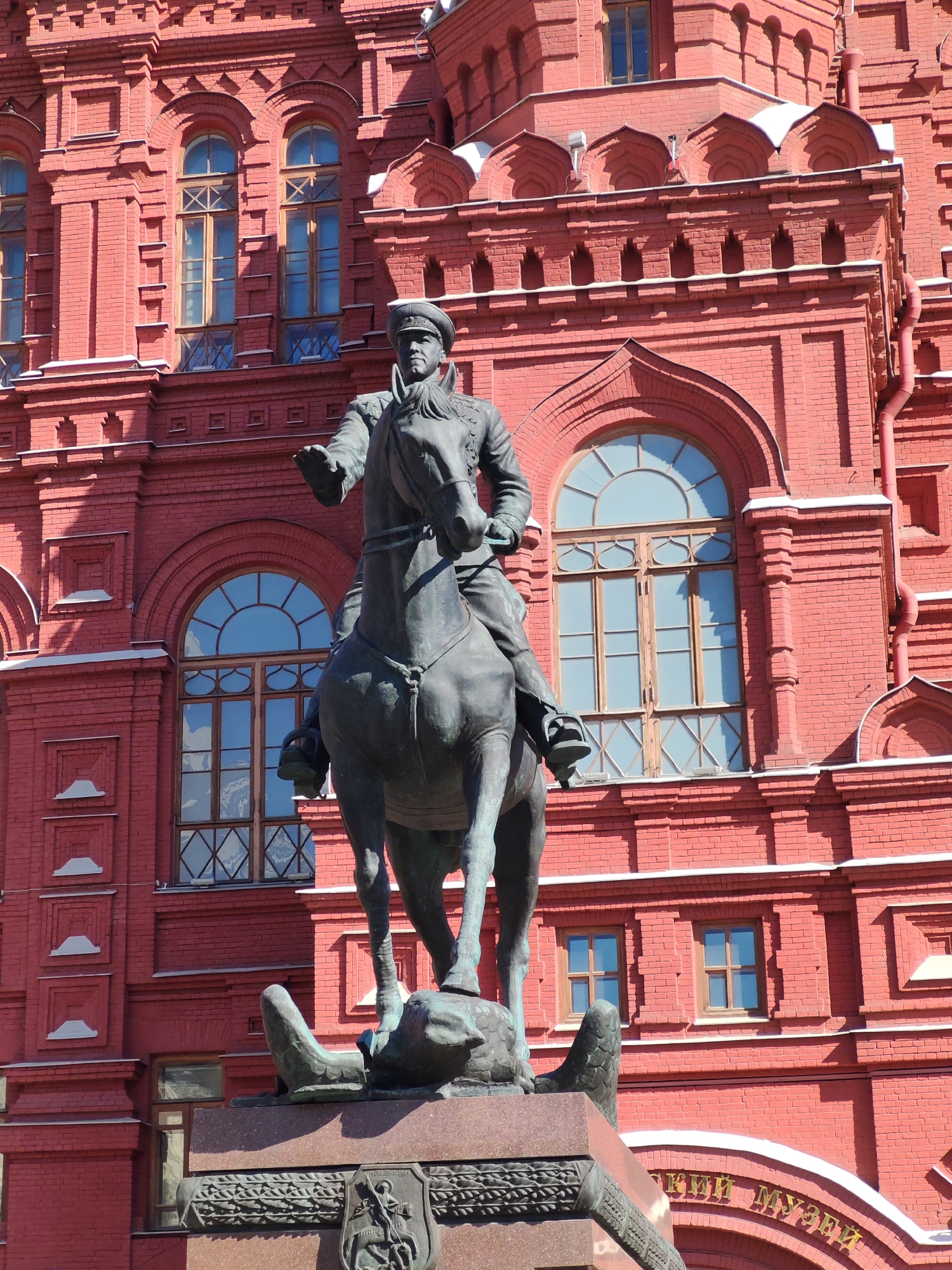 An outdoor, low-angle, full shot captures a bronze equestrian statue in front of a red brick building with arched windows and decorative brickwork. The statue depicts a man in a military uniform on a horse, with the man's left arm extended forward, pointing. The horse is rearing on its hind legs, standing on a large stone pedestal adorned with a relief and a coat of arms. The building behind is a multi-story structure with a textured red brick facade and numerous windows, some of which are arched and have diamond-shaped panes. Snow is visible on the roofline of the building. The overall scene suggests a historical or governmental location.