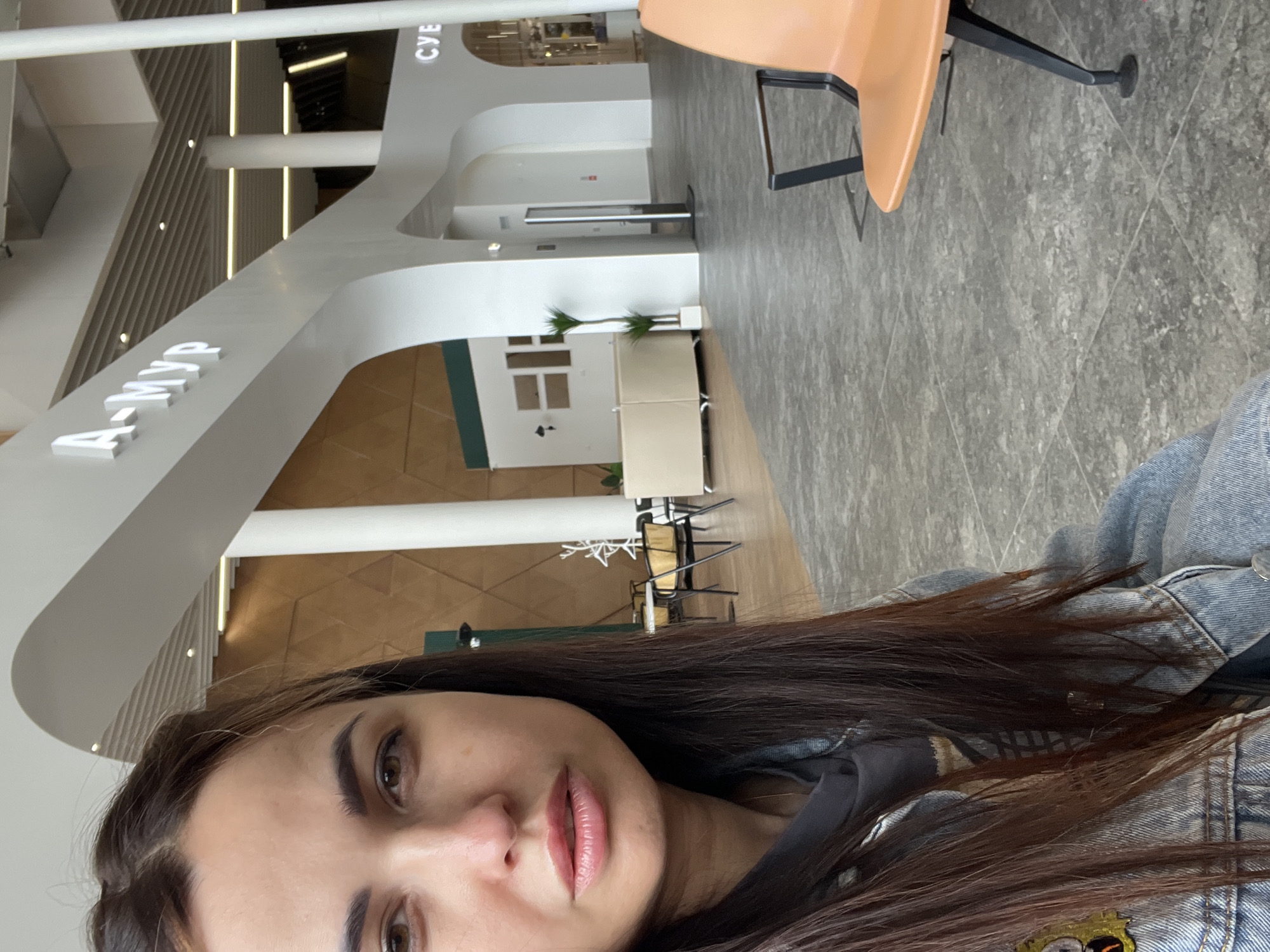A young woman with long, dark brown hair and brown eyes takes a selfie in what appears to be an indoor public space. She is wearing a denim jacket over a dark hoodie. Her lips are slightly parted, and she is looking directly at the camera. The background shows a modern architectural interior with white curved structures and signage. To the right, an orange chair is visible. The flooring is tiled. The overall impression is a candid shot taken while waiting or in transit. The location is identified as Unknown, Unknown.