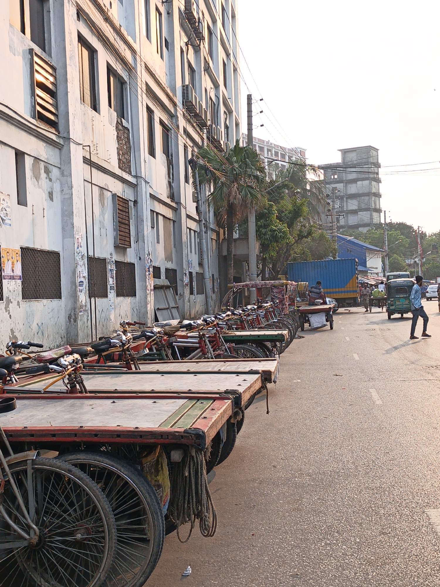The image depicts a street scene in Dhaka, Bangladesh. A row of cycle rickshaws is parked along the curb in front of a tall, weathered building with multiple windows, some covered with metal grates. A palm tree grows near the building. In the street, a blue and yellow truck and a green motorized rickshaw can be seen in the distance. A man in a light blue shirt and jeans walks along the right side of the road. The road itself is paved and shows signs of wear. A building in the distance appears to be under construction. Overhead, power lines crisscross the sky. The lighting suggests it is either early morning or late afternoon.
