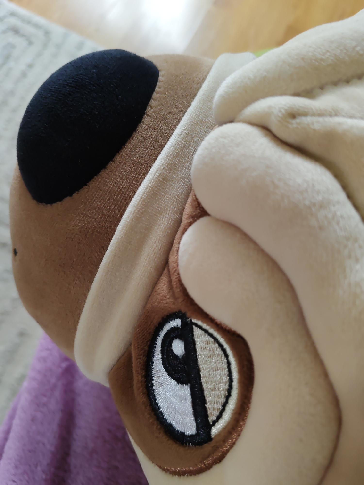 The image is a close-up, high-angle, shallow depth-of-field shot of a plush toy. The toy is a dog, with a black nose and brown and cream-colored fur. There is an embroidered patch on the side of the toy's face, which appears to be a stylized depiction of a dog's eye or a yin-yang symbol. The background is blurred and shows a light-colored surface that could be a carpet or rug, with a hint of wooden flooring. A purple fabric is visible in the lower left portion of the image. The image does not contain any information about the city of Olginskaya, Russia.