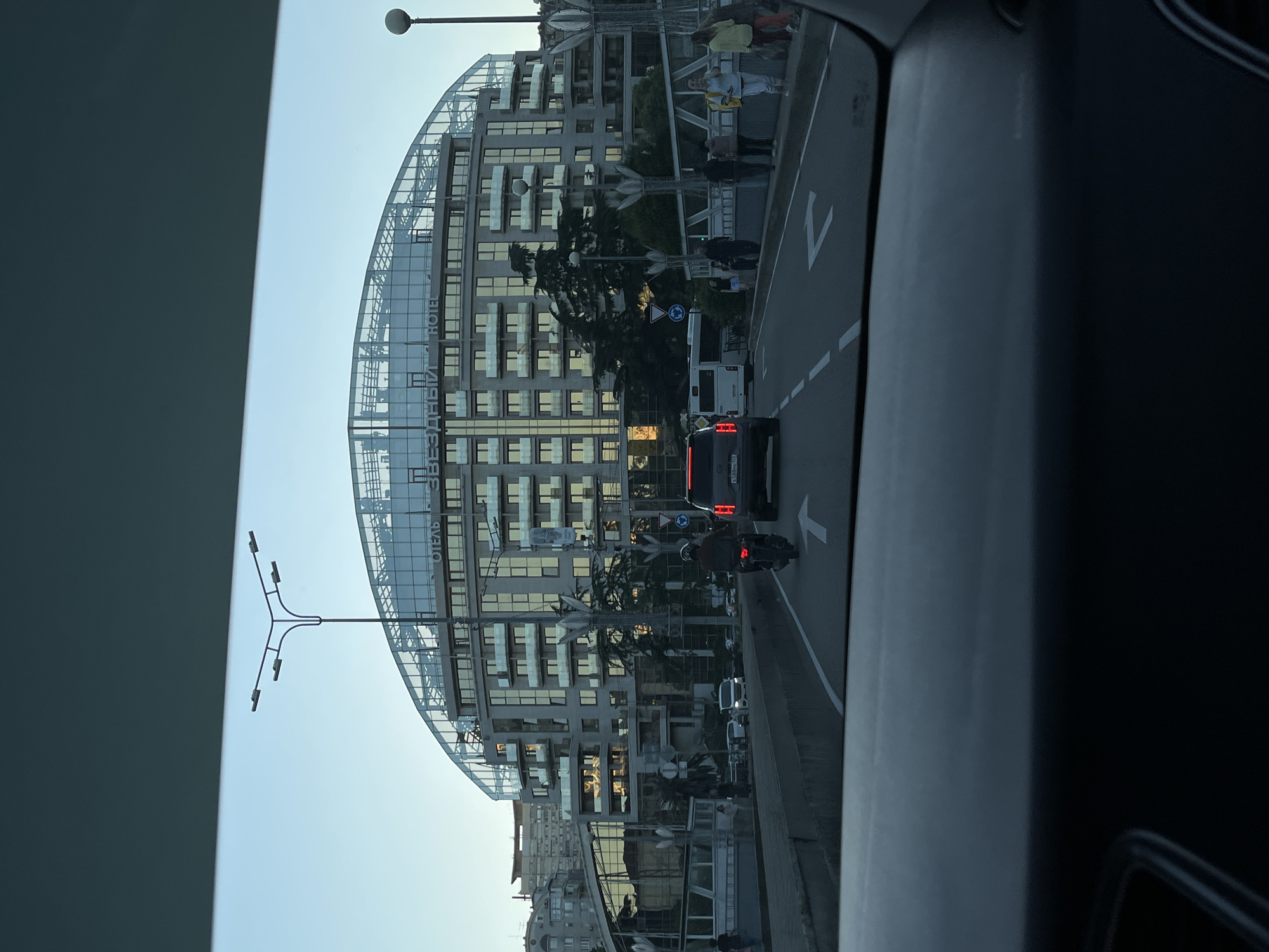 The image, taken from inside a vehicle, captures a street scene in Sochi, Russia, during what appears to be late afternoon or early evening. The prominent subject is a large, multi-story building with a distinctive curved, glass-paneled roof structure and numerous windows, some reflecting sunlight. Visible text on the building reads "ОТЕЛЬ ЗВЕЗДНЫЙ" (OTEL ZVEZDNY), identifying it as "Star Hotel."
In front of the building, a multi-lane road is visible with traffic. A dark SUV with Russian license plates (e.g., "P548MH 193") is in the foreground, followed by a white van or bus. A motorcyclist in a helmet is also on the road. Road markings include white arrows indicating direction and dashed lines. Several people are visible on a pedestrian area or sidewalk near the building and street.
Streetlights and traffic signs, including a circular blue sign (possibly a mandatory direction or roundabout sign) and a pedestrian crossing sign, are scattered along the roadside. Trees, possibly palm trees, are planted between the road and the building. The sky is clear and bright blue, suggesting fair weather. The scene depicts a busy urban environment.