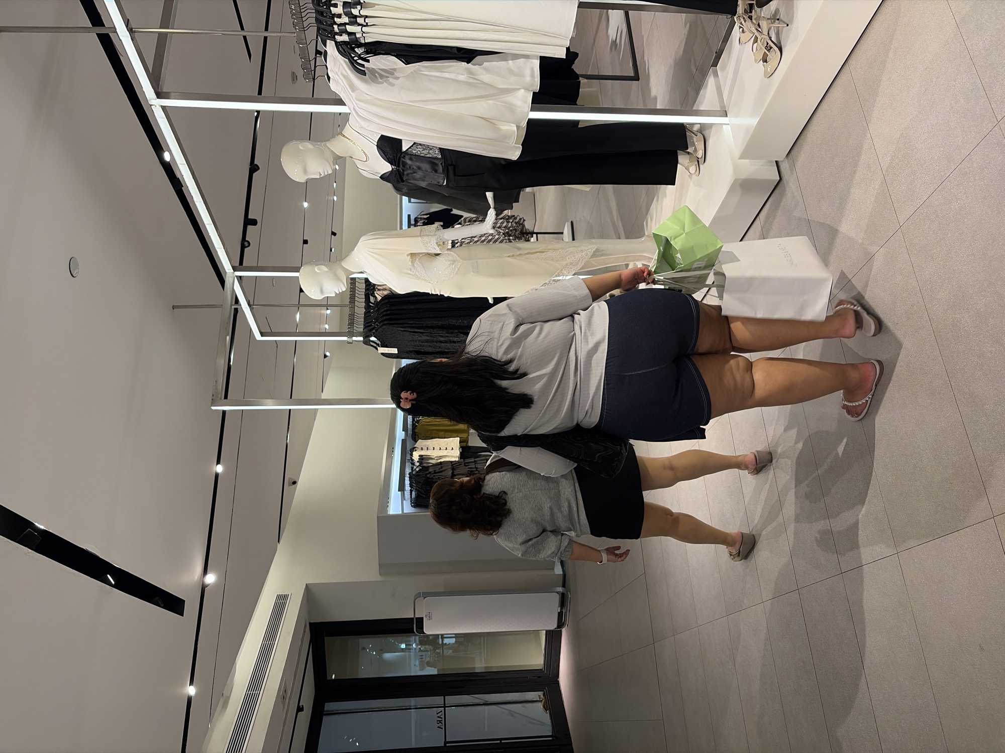 In Kuta, Indonesia, two women are walking through a brightly lit clothing store. The woman in front is wearing dark denim shorts and a light-colored, oversized short-sleeved shirt. She is carrying a white shopping bag in her right hand and a green paper bag in her left. She has dark hair pulled back with a pink clip. The woman behind her is wearing a grey long-sleeved shirt and a black mini-skirt. She has dark, curly hair and is carrying a black tote bag over her left shoulder. They are walking away from the camera, towards mannequins displaying clothing. The store has a tiled floor and modern light fixtures.