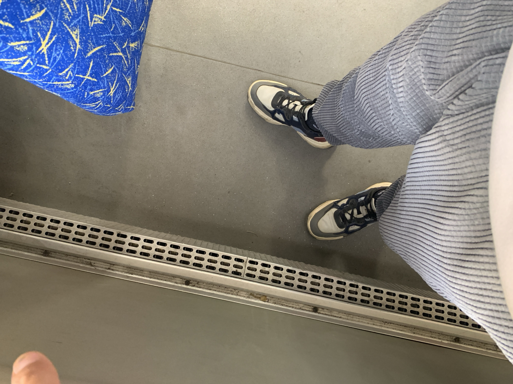 The image shows a person's legs and feet standing on a gray tiled floor. The person is wearing light blue corduroy pants with a pinstripe pattern and sneakers. The sneakers are a mix of white, gray, and dark blue. To the left of the person is a metallic ventilation grate with a series of rectangular openings, and beyond that, a smooth, light gray surface, possibly a wall or partition. To the right, a portion of a blue seat with a yellow and blue abstract pattern is visible. The scene appears to be indoors, possibly in a mode of public transportation or a waiting area.