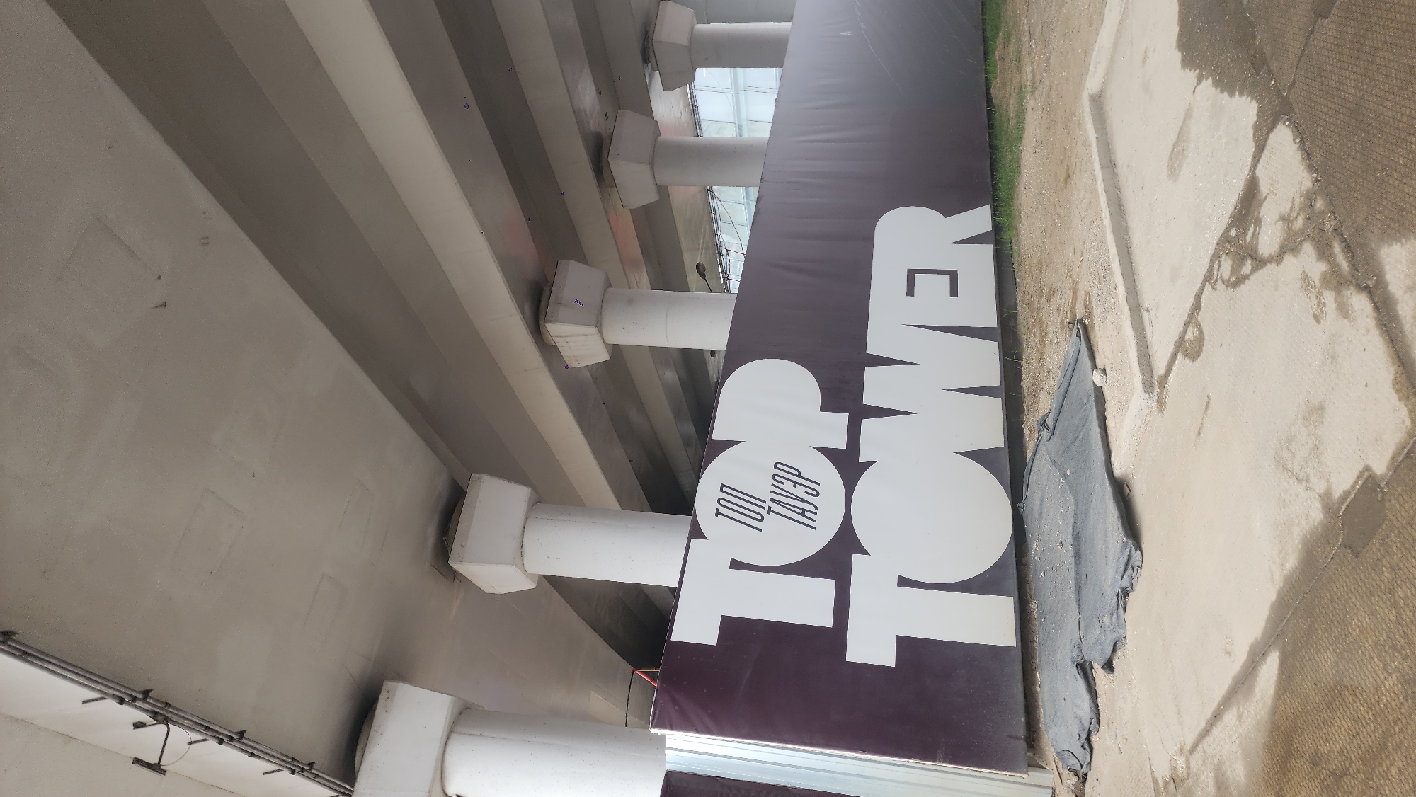The image shows a sign for "Top Tower" in Moscow, Russia. The sign is brown with white lettering and is located under a bridge or overpass. The area around the sign is paved, with some dirt and vegetation visible. No people are visible in the image.