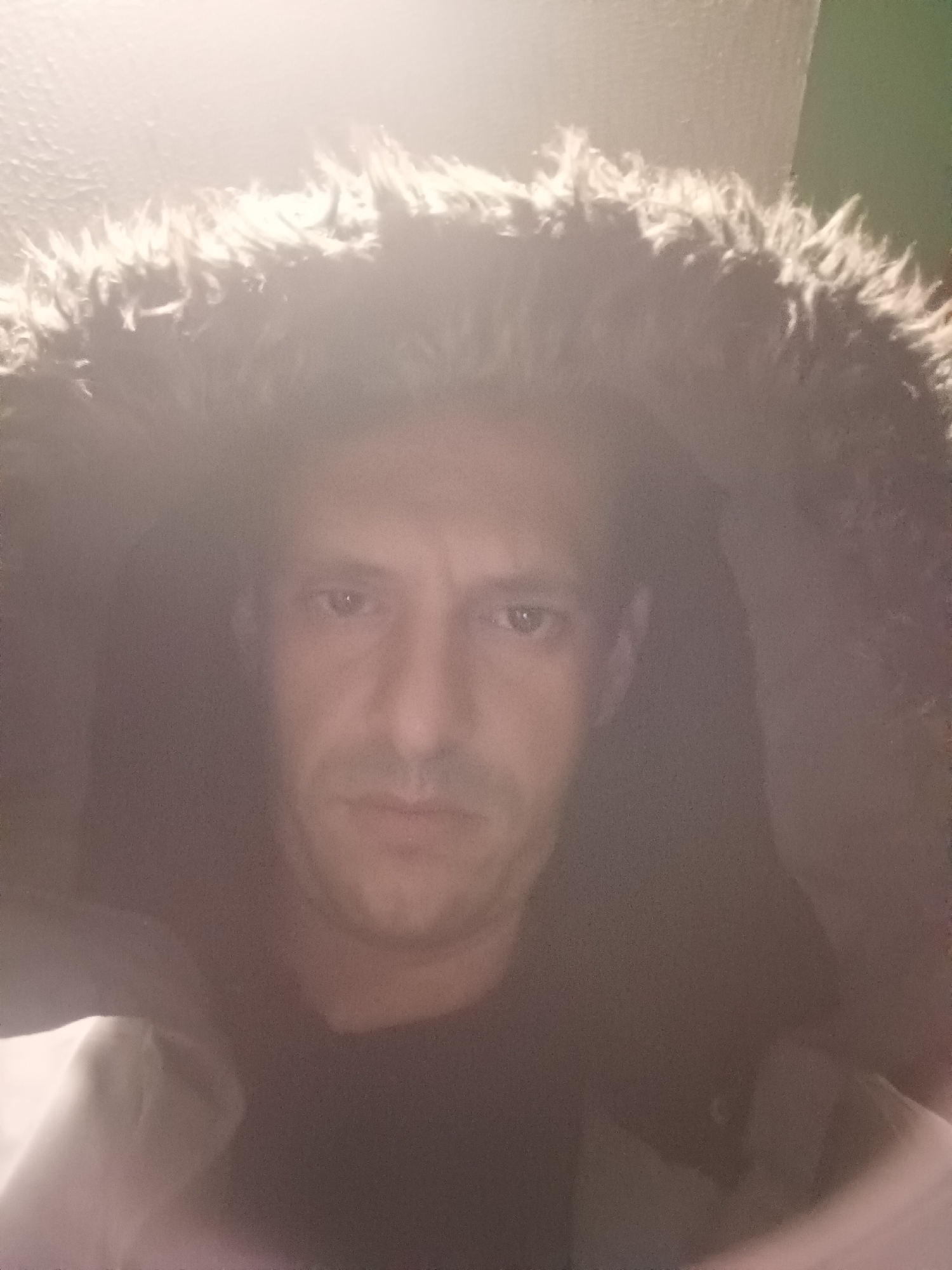 This is a portrait of an adult male in Burgas, Bulgaria. The subject is depicted from the chest up, positioned centrally, looking directly at the viewer with a neutral expression. He is wearing a dark-colored garment under a light-colored jacket or coat featuring a prominent, shaggy, light-colored faux fur-lined hood that frames his face. The upper portion of the image, including the fur hood and overhead area, exhibits significant overexposure and glare from a bright light source. The background is dark and indistinct, appearing to be an indoor environment, with a subtle greenish hue visible on the upper right side. The image quality is somewhat soft and shows evidence of lens flare or reflections.