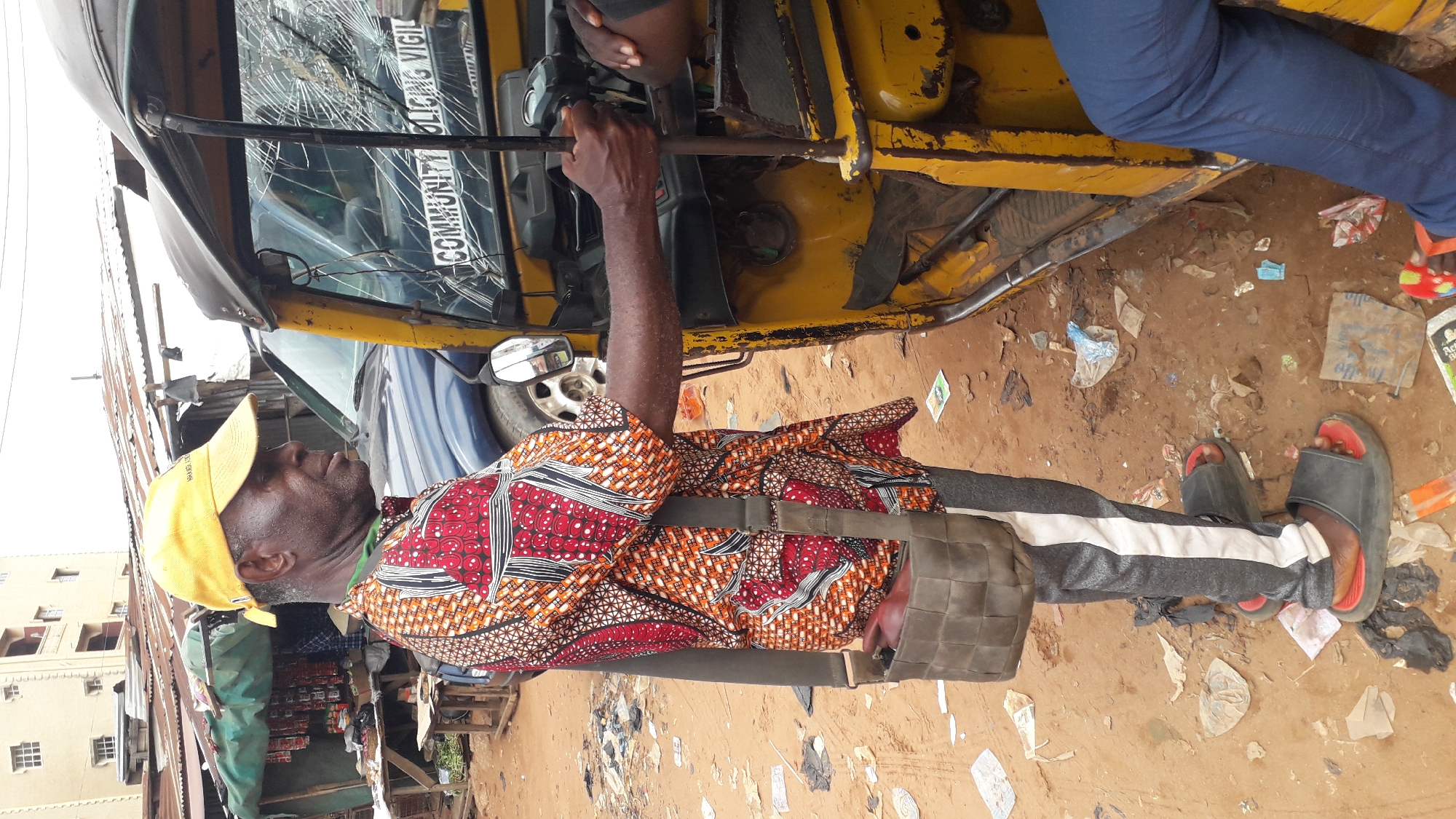 The image shows a man in Nkpor, Nigeria, standing next to a yellow three-wheeled vehicle with a cracked windshield labeled "COMMUNITY POLICING VIGIL." He is wearing a patterned shirt, grey pants with a white stripe, and black sandals. He also has a yellow hat and a bag slung across his shoulder. He is holding onto the vehicle's handlebars. The ground is dirt with scattered trash. A building and a shop are visible in the background.
