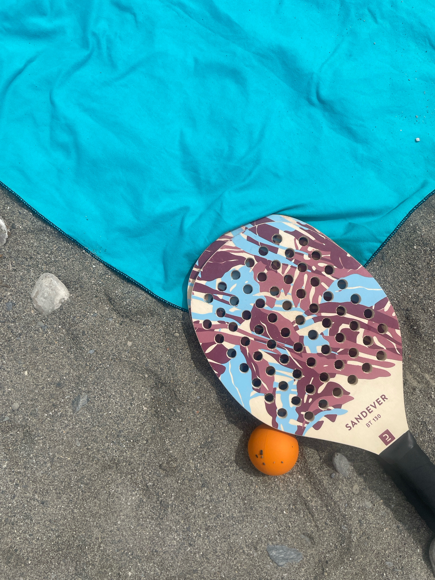 A beach paddleball set rests on the sandy shore in La Herradura, Spain. A bright turquoise towel is spread out to the upper left, with its edge forming a diagonal line across the sand. A paddleball racket with a camouflage pattern of purple, blue, and beige is positioned in the lower right of the frame, angled slightly upwards. Beside the paddle, a small, bright orange ball sits on the sand. The sand is speckled with small grey pebbles and darker sand particles. The image is taken from a high-angle perspective, looking down onto the scene.