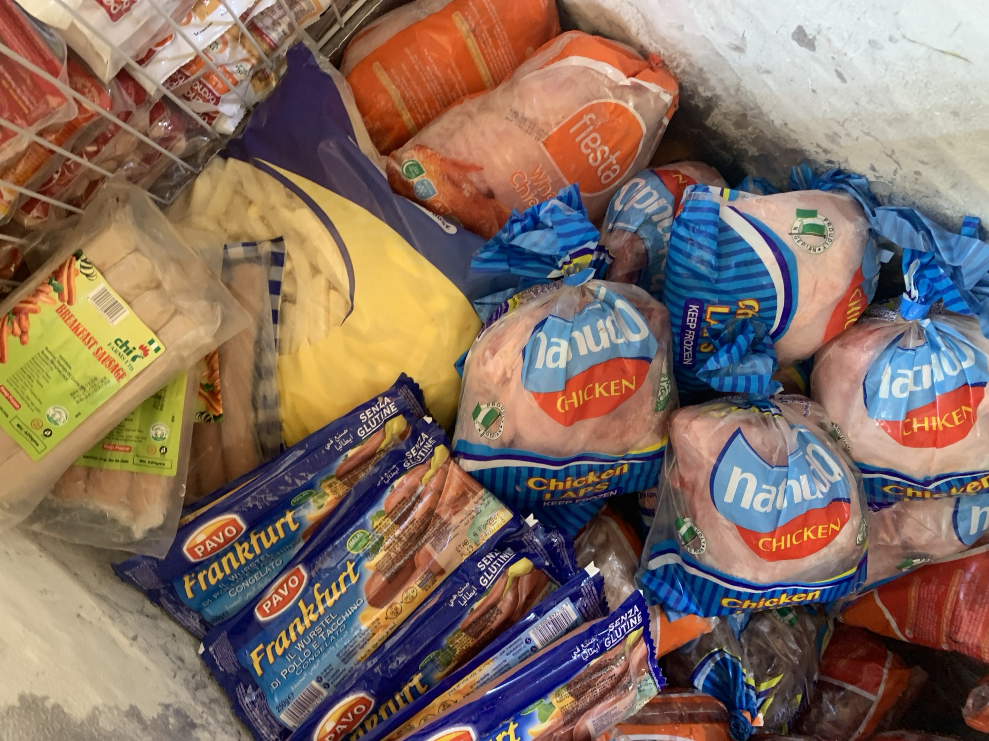 In an indoor frozen food display, multiple packages of frozen food are visible. In the foreground and left, several blue and yellow packages of "Frankfurt" sausages are stacked. To the right of the sausages, several clear plastic bags contain whole chickens, some with "Fiesta" branding and others with "Nani" branding. In the background, additional packages of frozen food are visible, including a large yellow bag and a package of breakfast sausages. The environment appears to be a commercial freezer or display case, with a frosty interior. No people or specific actions are depicted.