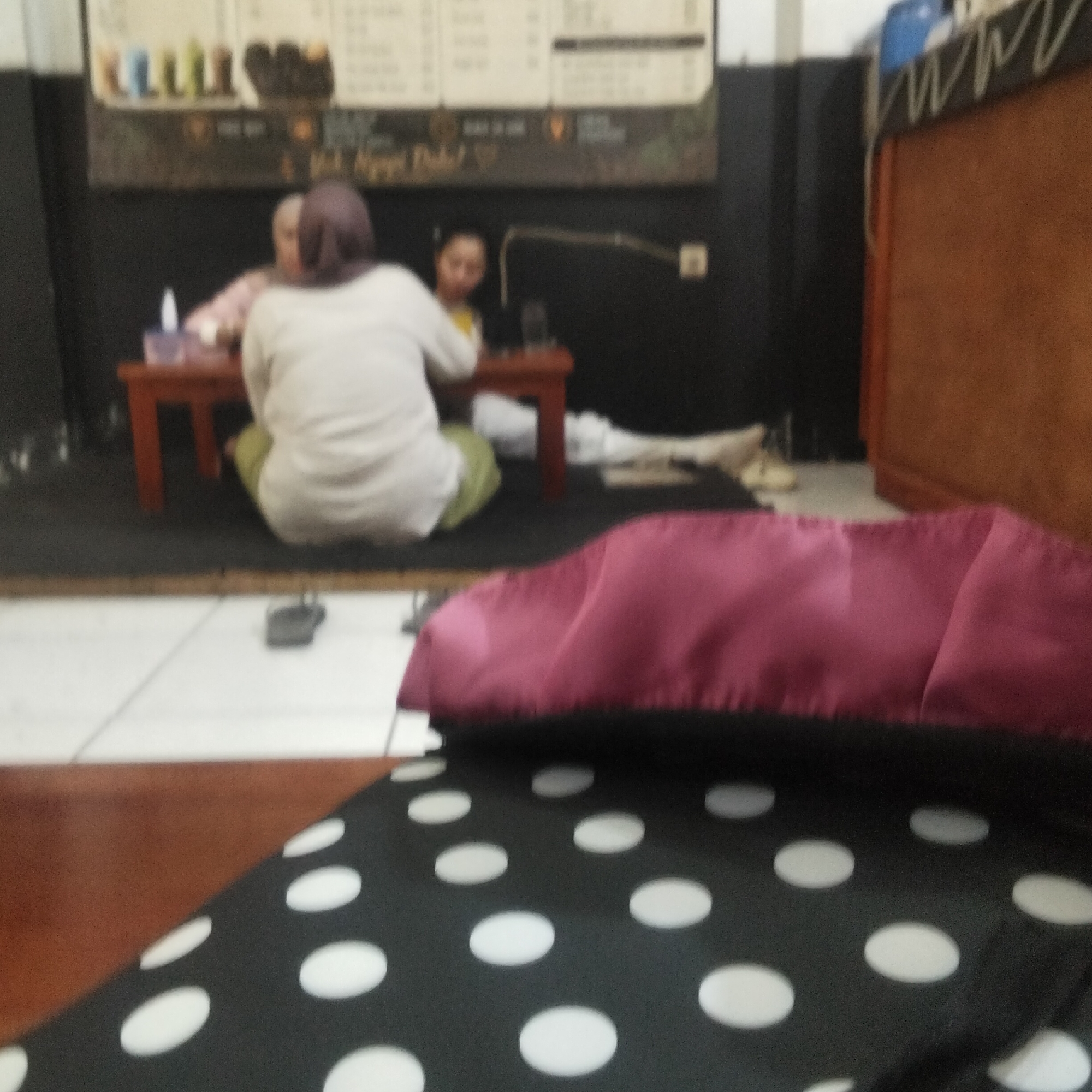 The image depicts an indoor scene, likely a cafe or restaurant in Sukamanah, Indonesia. In the background, a menu board with text and images of drinks is visible. Two individuals are seated at a low, reddish-brown table. One person, wearing a light-colored top and a purple headscarf, is facing away from the camera. Another person, in a yellow top and a dark head covering, is partially visible and appears to be interacting with the first individual. On the table, there are some containers, possibly for condiments or drinks.
In the foreground, a plush, dark fabric with white polka dots is spread out, and a burgundy fabric is draped over it. The floor appears to be tiled with white squares. The overall lighting suggests an indoor environment, possibly with ambient or artificial light sources. The image has a slightly blurred quality, typical of a low-light or distance shot. The scene conveys a casual, relaxed atmosphere.
