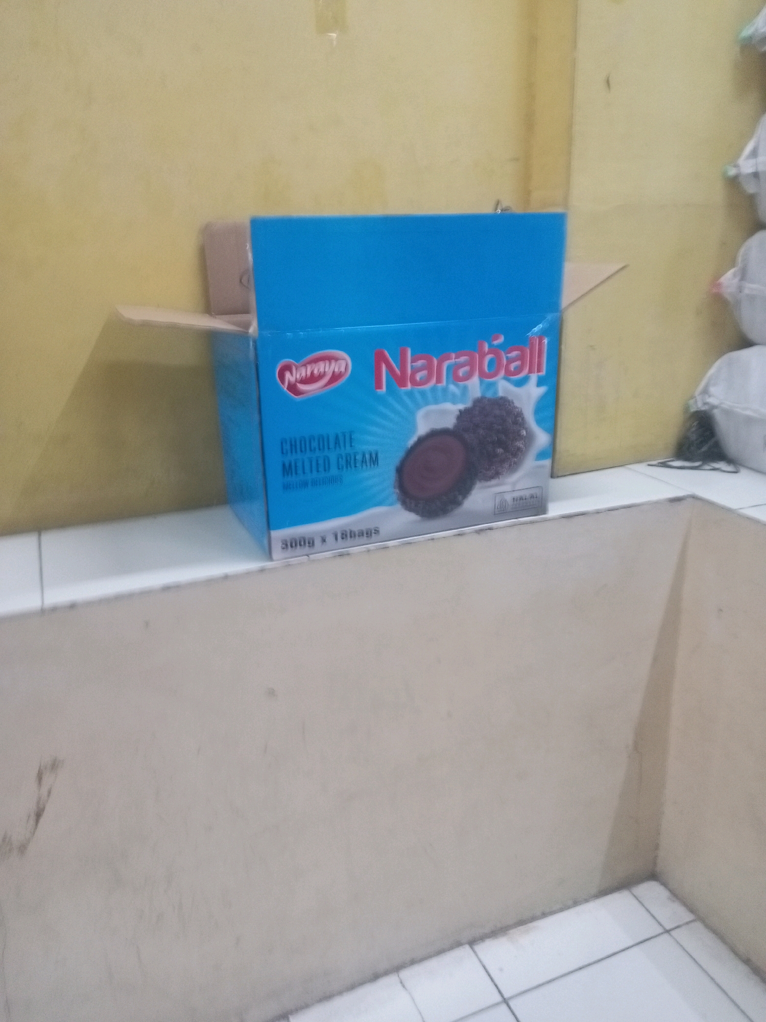 The image shows a cardboard box of "Naraya Naraball Chocolate Melted Cream" snacks. The box is blue and open, revealing its contents. Two chocolate snacks with a creamy filling are depicted on the front of the box, along with a "HALAL" logo. The box is placed on a white surface, possibly a table or shelf, in front of a yellow wall. In the background, to the right, a stack of white bags is visible. The scene appears to be an indoor setting, likely a storage area or shop in Batam, Indonesia.