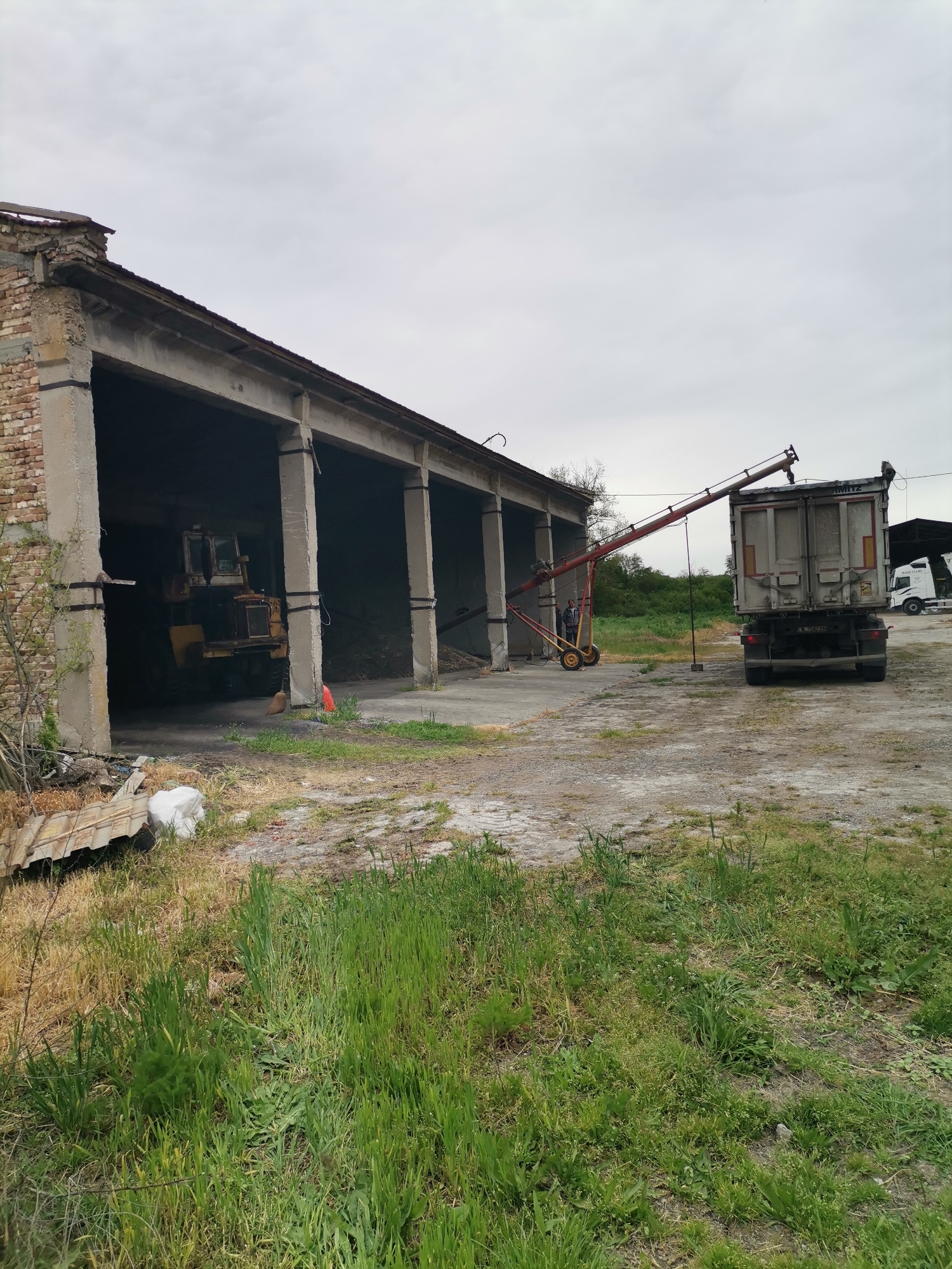 The image depicts a rural scene in what appears to be an agricultural or industrial setting, potentially in or near Kochovo, Bulgaria. The foreground consists of vegetation transitioning from dry, patchy ground to taller, green grass.
To the left stands a dilapidated, open-sided structure with brick and concrete pillars supporting a roof. Inside, a yellow bulldozer is partially visible. Debris and discarded items are scattered near the entrance of the structure.
A truck with a metal container bed is parked in the center of the scene, connected to a red and yellow auger or conveyor system. Two figures are standing near the auger, appearing to be involved in the process of loading or unloading materials into or from the truck. A support brace is connected to the ground.
In the background, a white tractor is visible next to a black covered shed. The overall setting is overcast with a grey sky. The ground surrounding the buildings is a mix of concrete and bare earth.