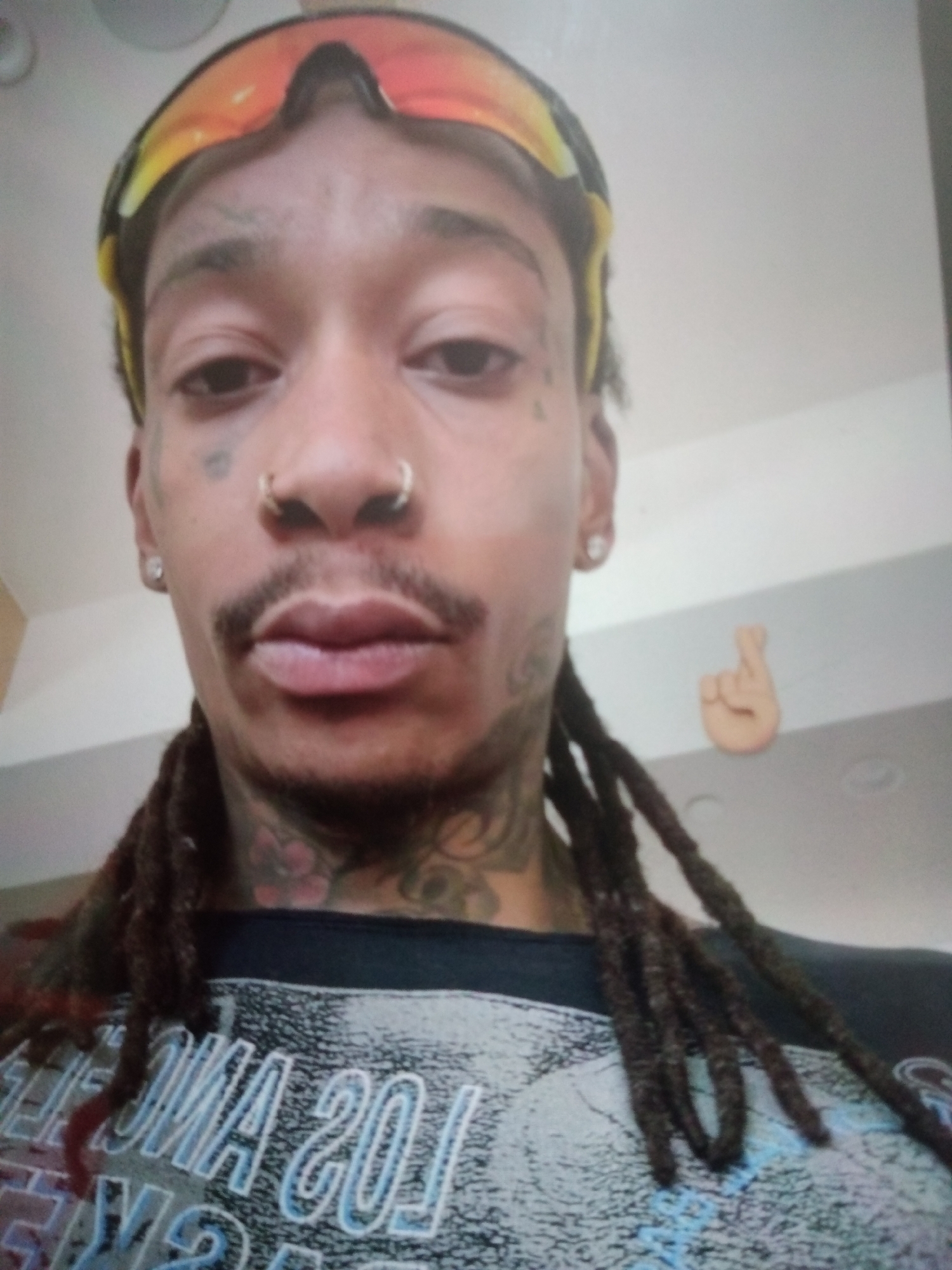 The image is a close-up, selfie-style portrait of a man with dreadlocks, wearing sunglasses pushed up onto his forehead. He has a septum piercing and a tattoo on his neck. He is wearing a dark t-shirt with white and blue lettering. A small, peach-colored figurine of a hand with crossed fingers is visible on the wall behind him. The background appears to be an indoor setting with a plain white ceiling and wall. There is no visible indication of the image being taken in Jalingo, Nigeria.