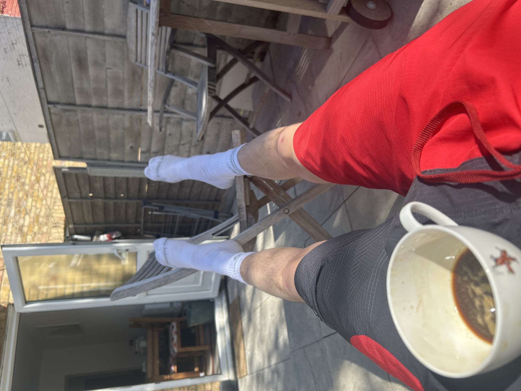 A person is sitting on a wooden chair outdoors with their legs extended and wearing white socks, red shorts on the right leg, and black shorts on the left leg. The person is holding a white mug with coffee in it. The background shows a wooden fence, a table and chair, and a building with a glass door and windows. The location cannot be identified from the image alone.