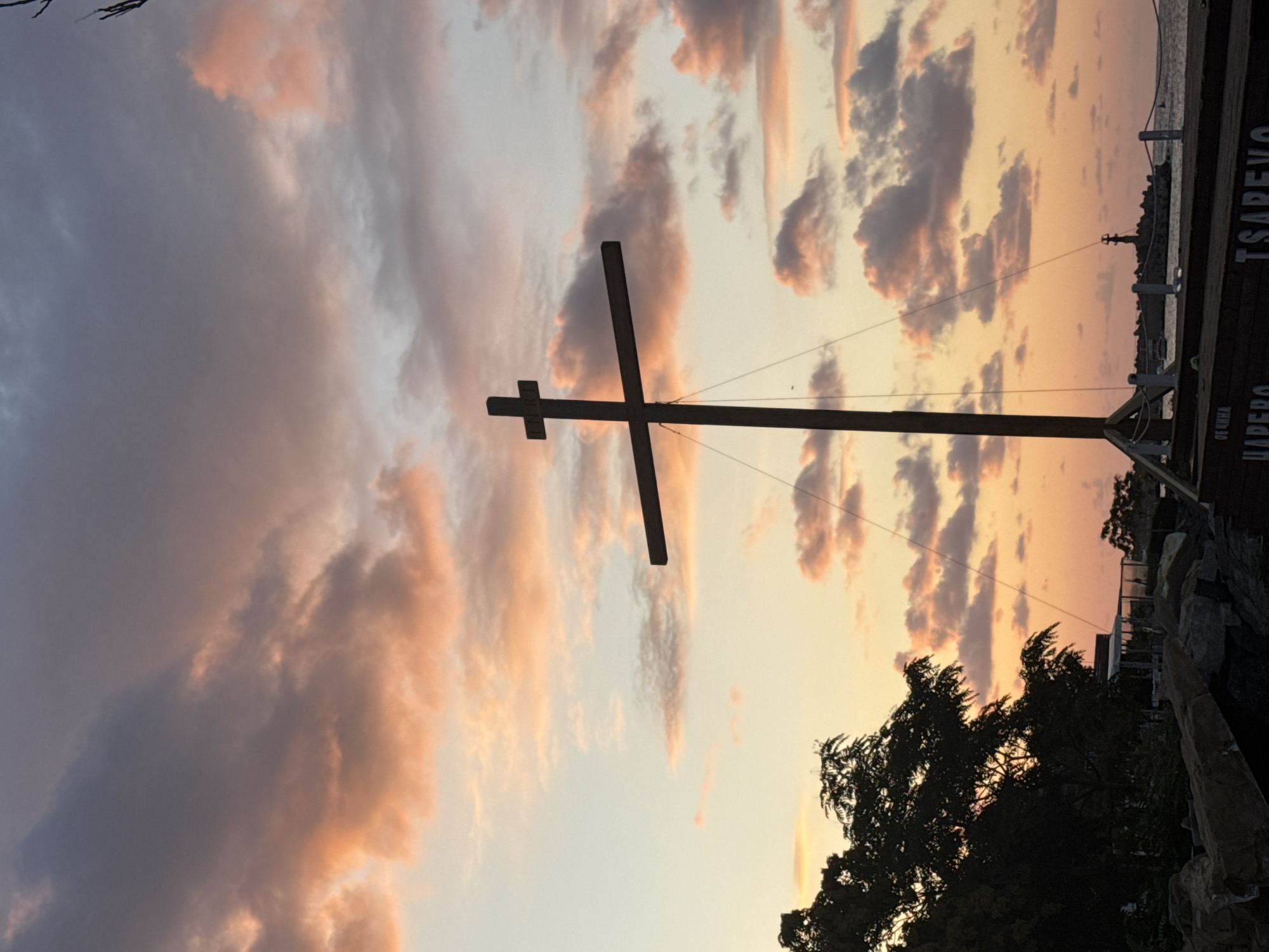 This image captures a sunset scene in Tsarevo, Bulgaria. The foreground is dominated by a large wooden cross, with the letters "INRI" visible on its horizontal beam. The cross stands against a backdrop of a colorful sky filled with pink, orange, and gray clouds, illuminated by the setting sun. To the left of the cross, silhouetted trees and rocky terrain are visible. In the distance, a lighthouse stands on a breakwater, with the sea extending to the horizon. The overall impression is one of serene, natural beauty with a religious motif.