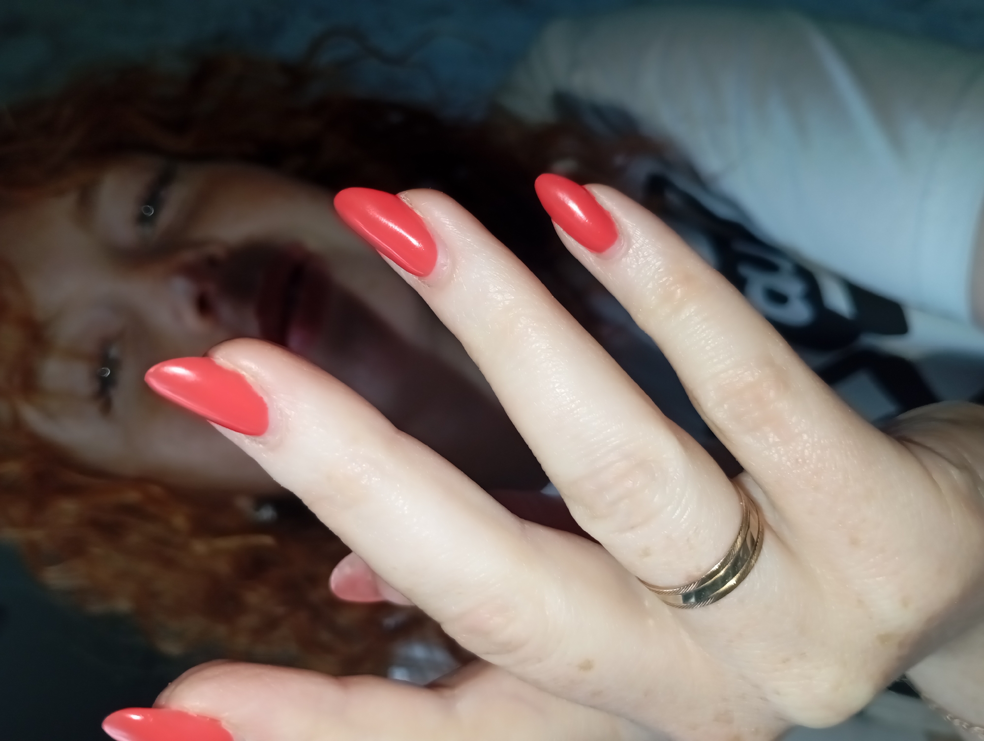 A photograph depicts a woman with fair skin, red hair, and a visible lip piercing. She is wearing a white T-shirt with black print and her right hand is prominently displayed in the foreground. Her nails are painted a bright red color, and a gold-colored ring adorns her ring finger. The focus is on the hand and the woman's face is blurred in the background. There is no identifiable evidence in the image to confirm it was taken in Yurgamysh, Russia.